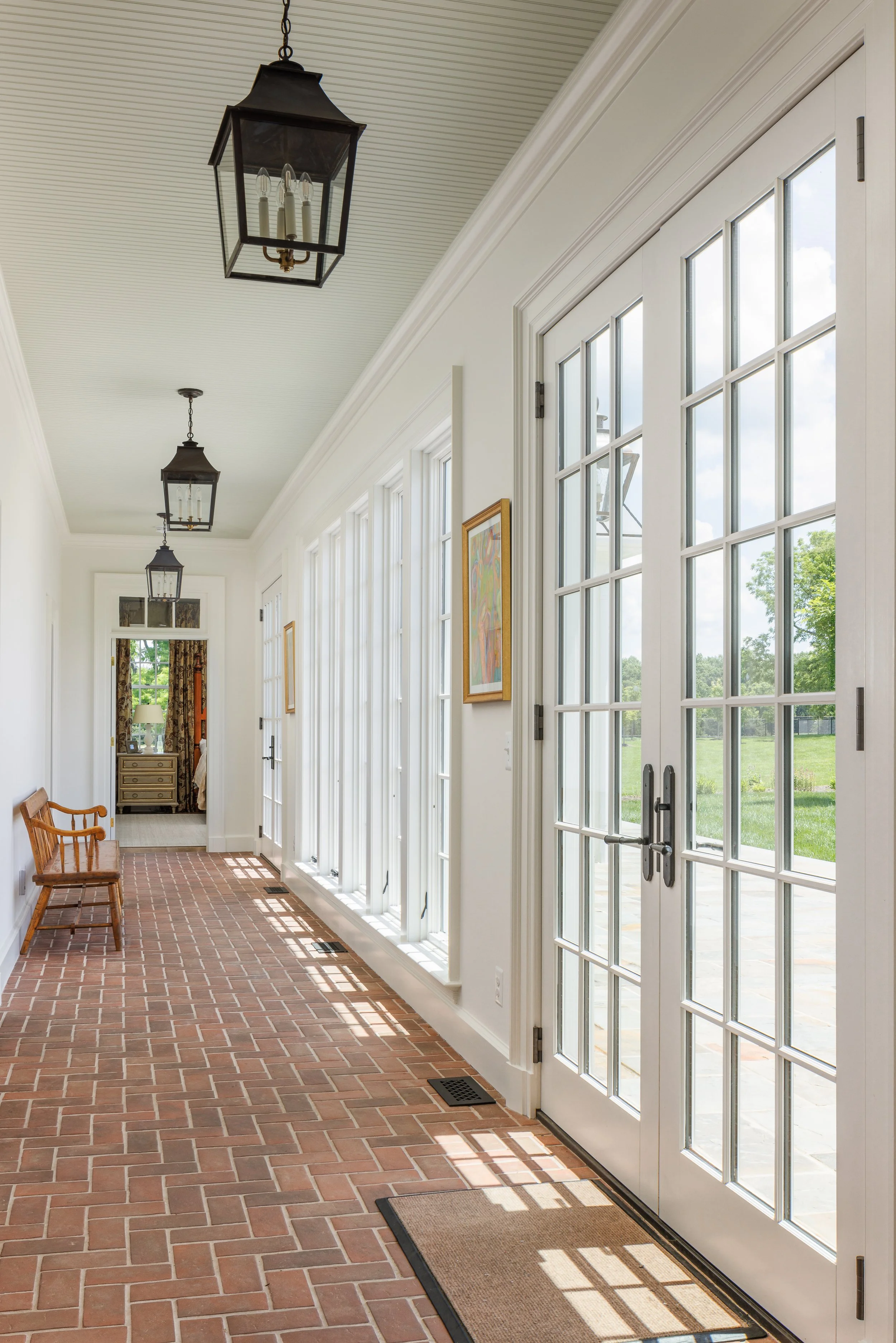 Bright indoor hallway with brick flooring, white walls, large glass-paneled doors, and ceiling lanterns. A wooden bench is on the left, and artwork decorates the walls.