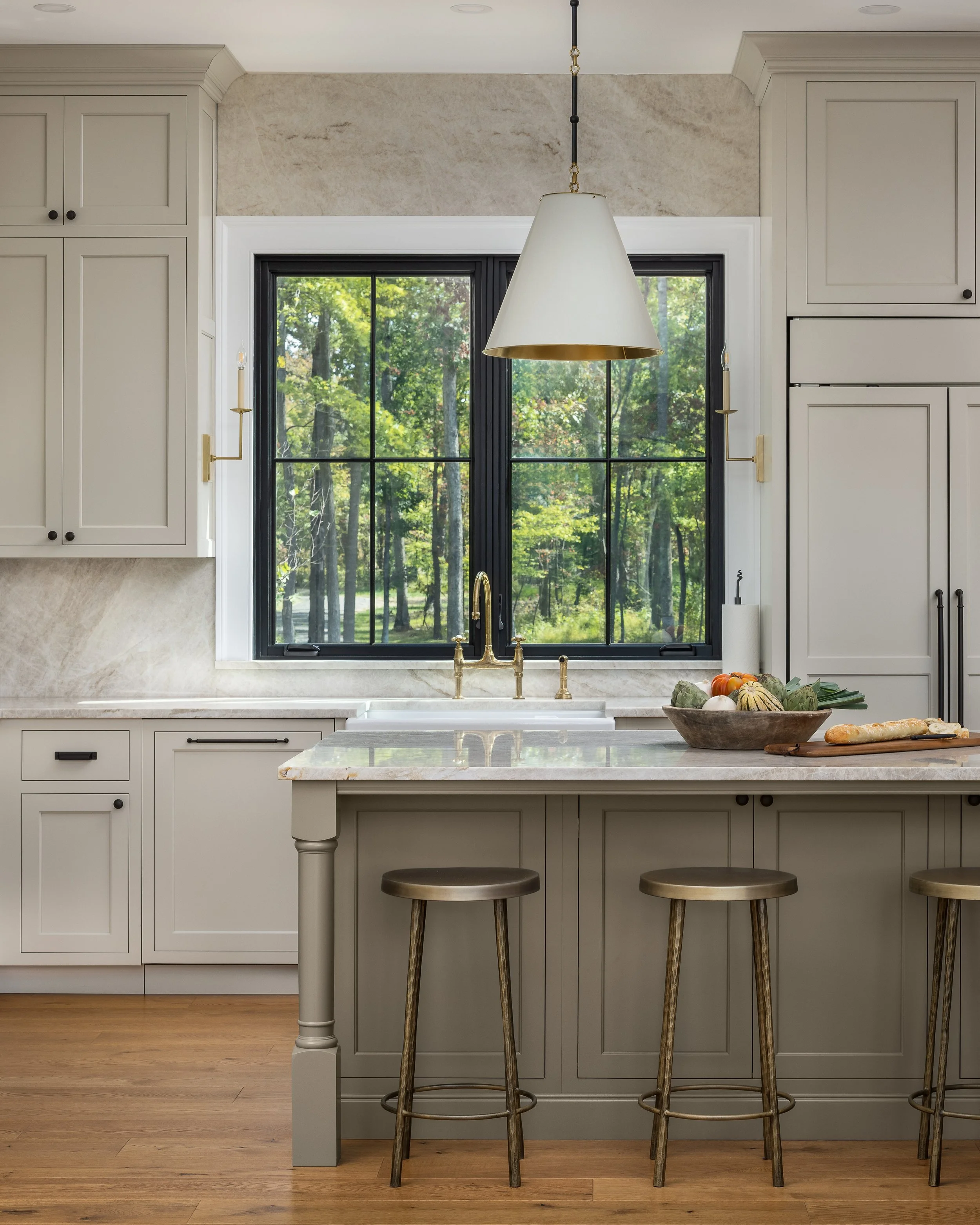 Modern kitchen with white cabinets, a large window showing a green outdoor scene, a white marble island with three gold bar stools, a bowl of vegetables on the island, a white sink under the window, and a pendant light hanging above.