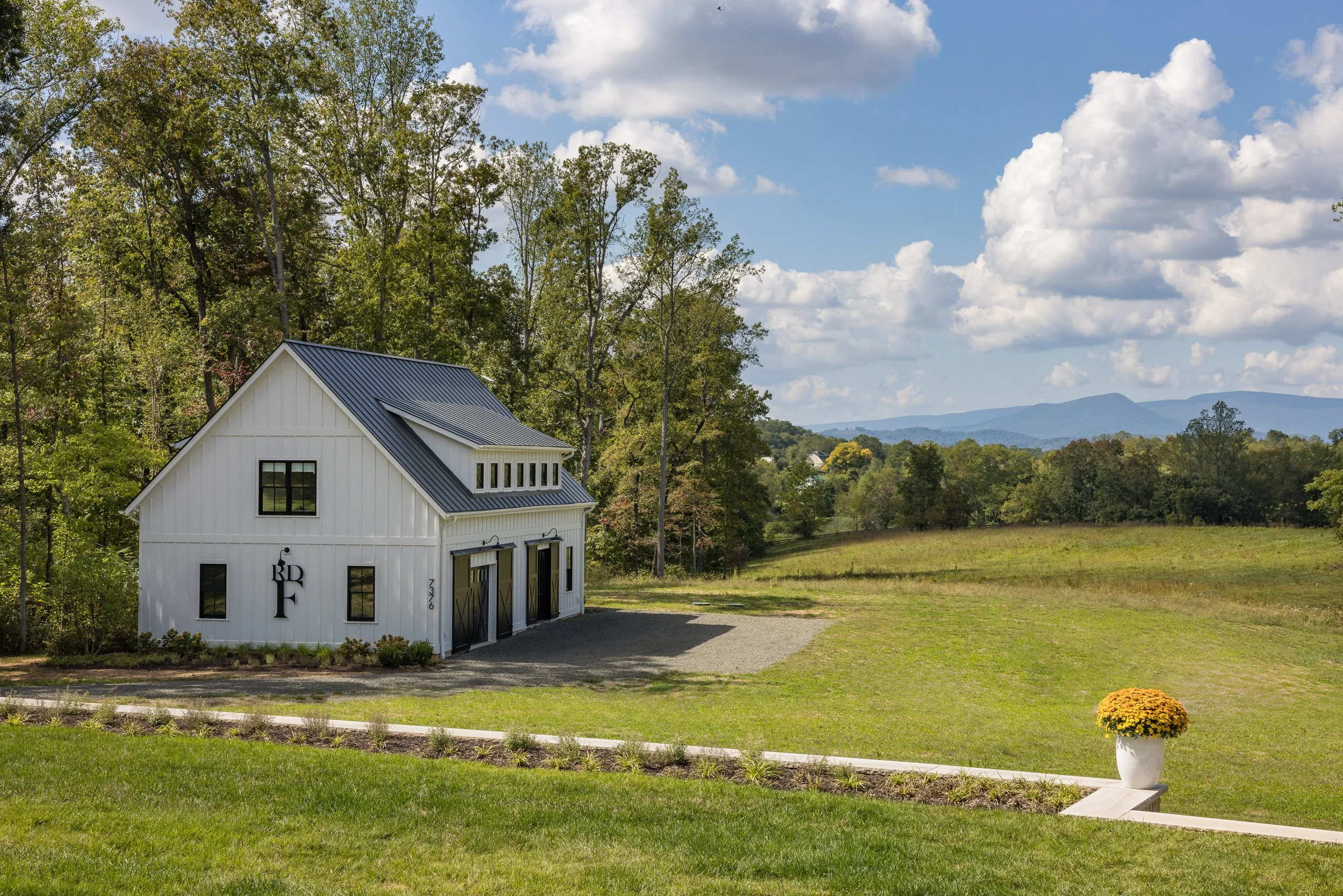 A white barn with black doors and roof, situated in a lush green landscape with trees and distant mountains under a partly cloudy sky.