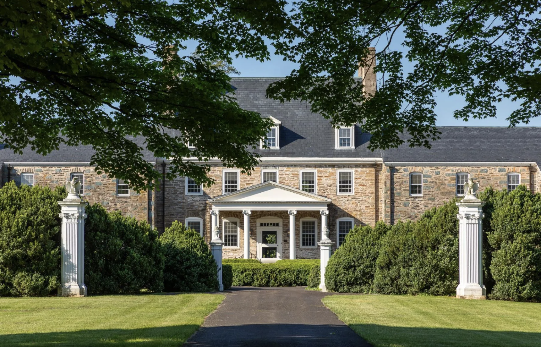 A large stone historic house with white columns at the entrance, surrounded by greenery and a paved driveway, under a clear blue sky.