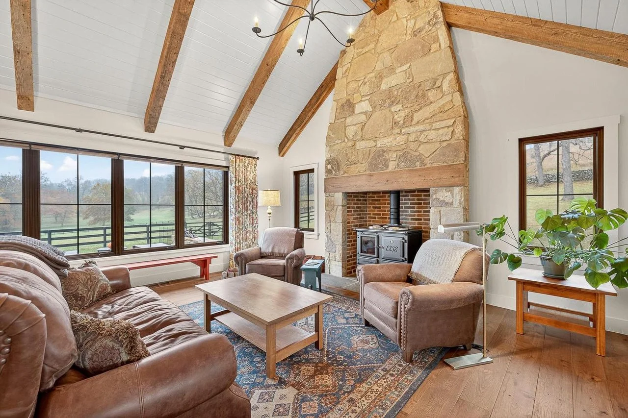 Cozy living room with a large stone fireplace, wooden beams on the ceiling, leather and fabric armchairs, a wooden coffee table, windows revealing a scenic outdoor view, a patterned rug, a potted plant, and a side table with a lamp.