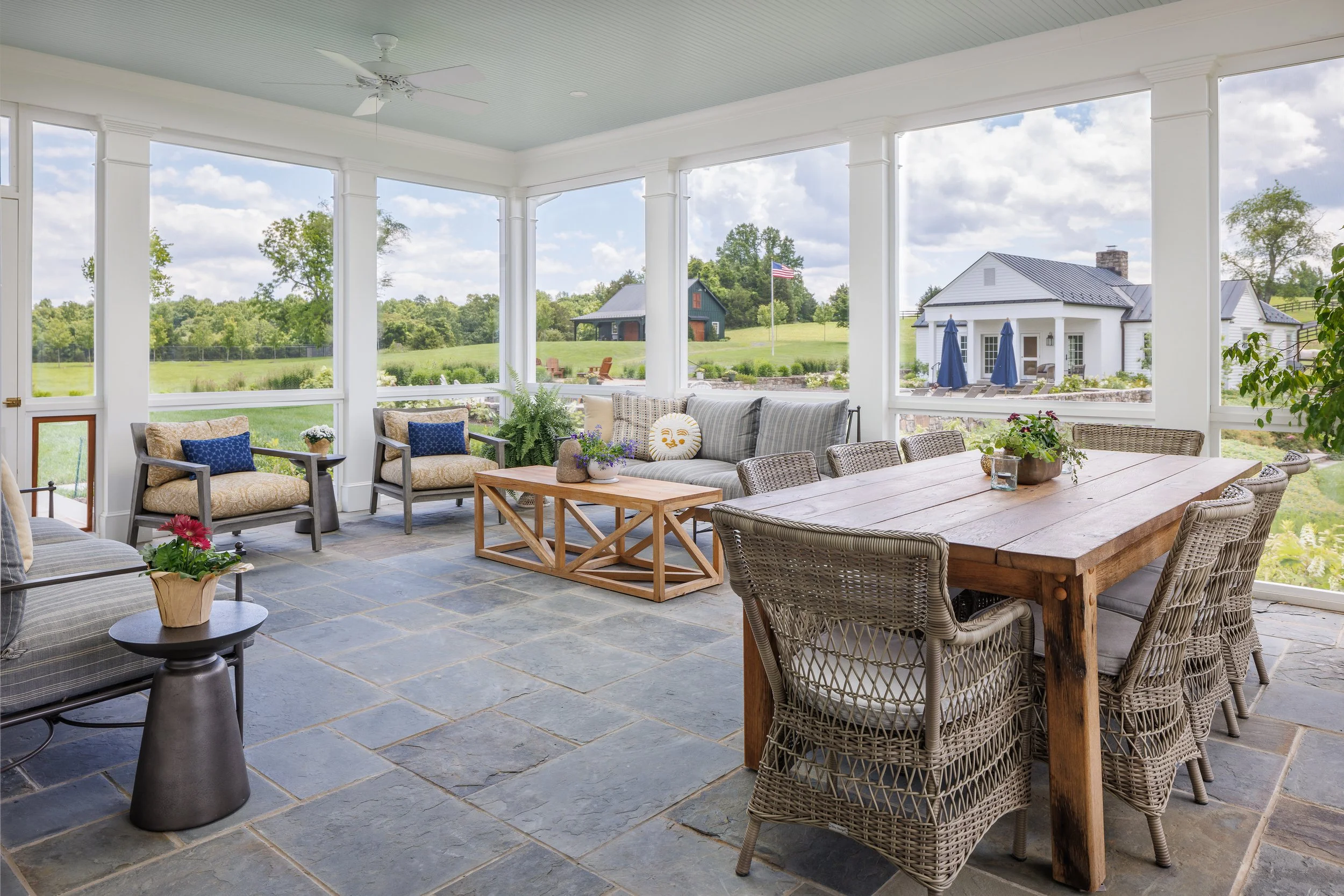 Bright porch or sunroom with large windows showing a green lawn and trees outside. Contains a wooden dining table with eight woven chairs, a long cushioned sofa, two armchairs with blue pillows, and a small coffee table with plants. The floor is ston