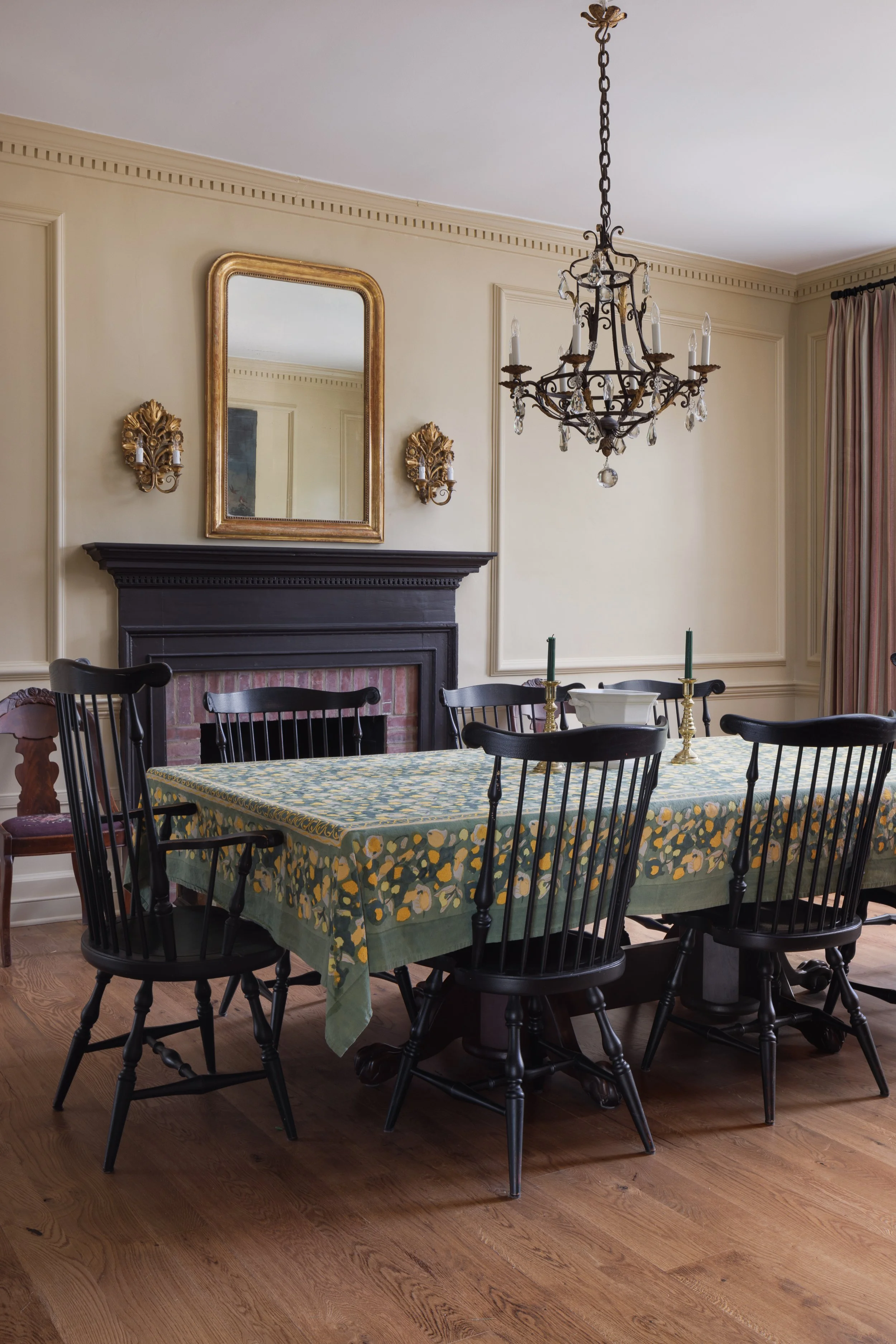 A dining room with a table covered with a floral tablecloth, black chairs, a fireplace with a mirror above it, gold wall sconces, a chandelier, and candles on the table.