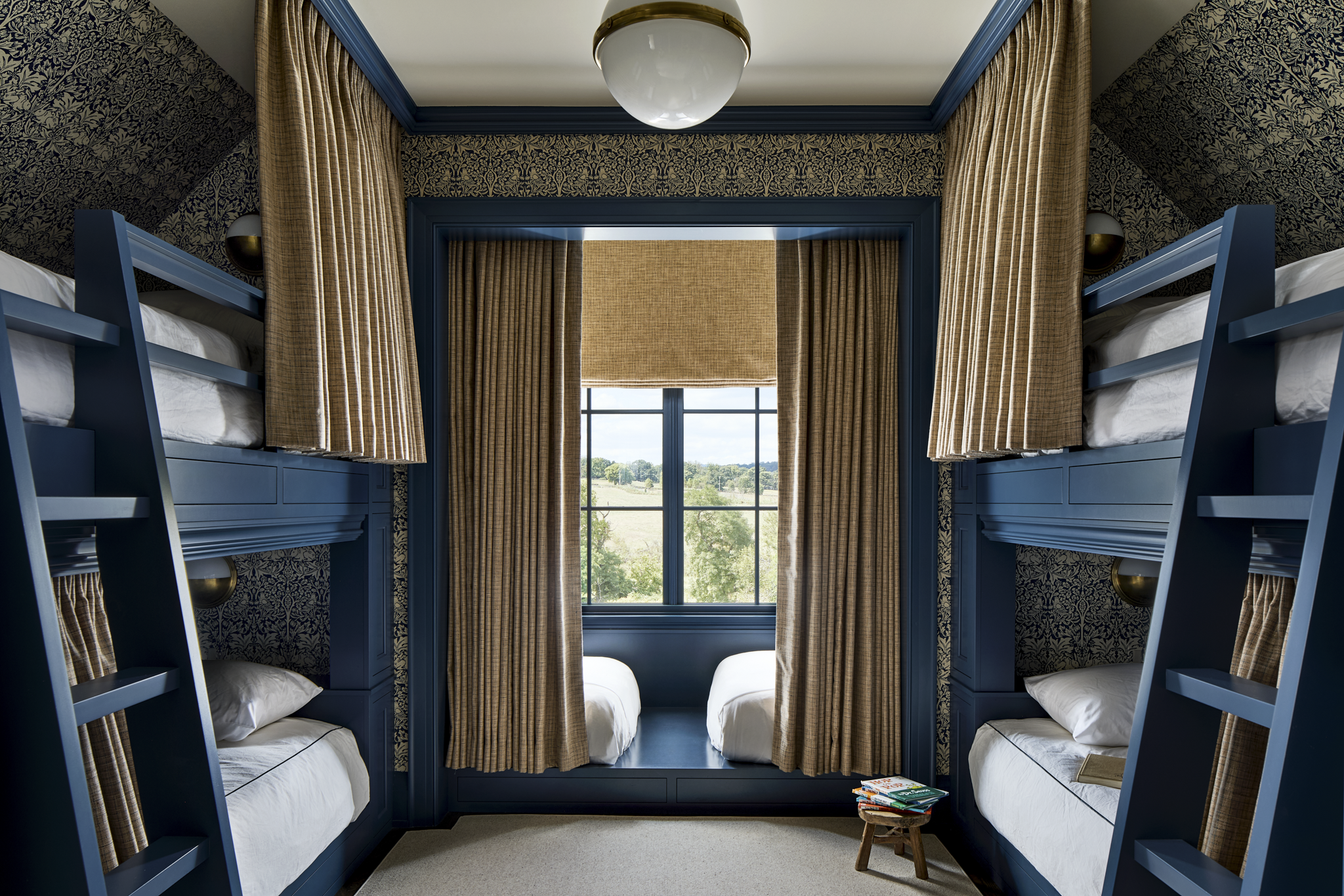 A room with bunk beds on each side, blue wood frames, beige curtains, a large window with a scenic view, and a small stool with magazines.