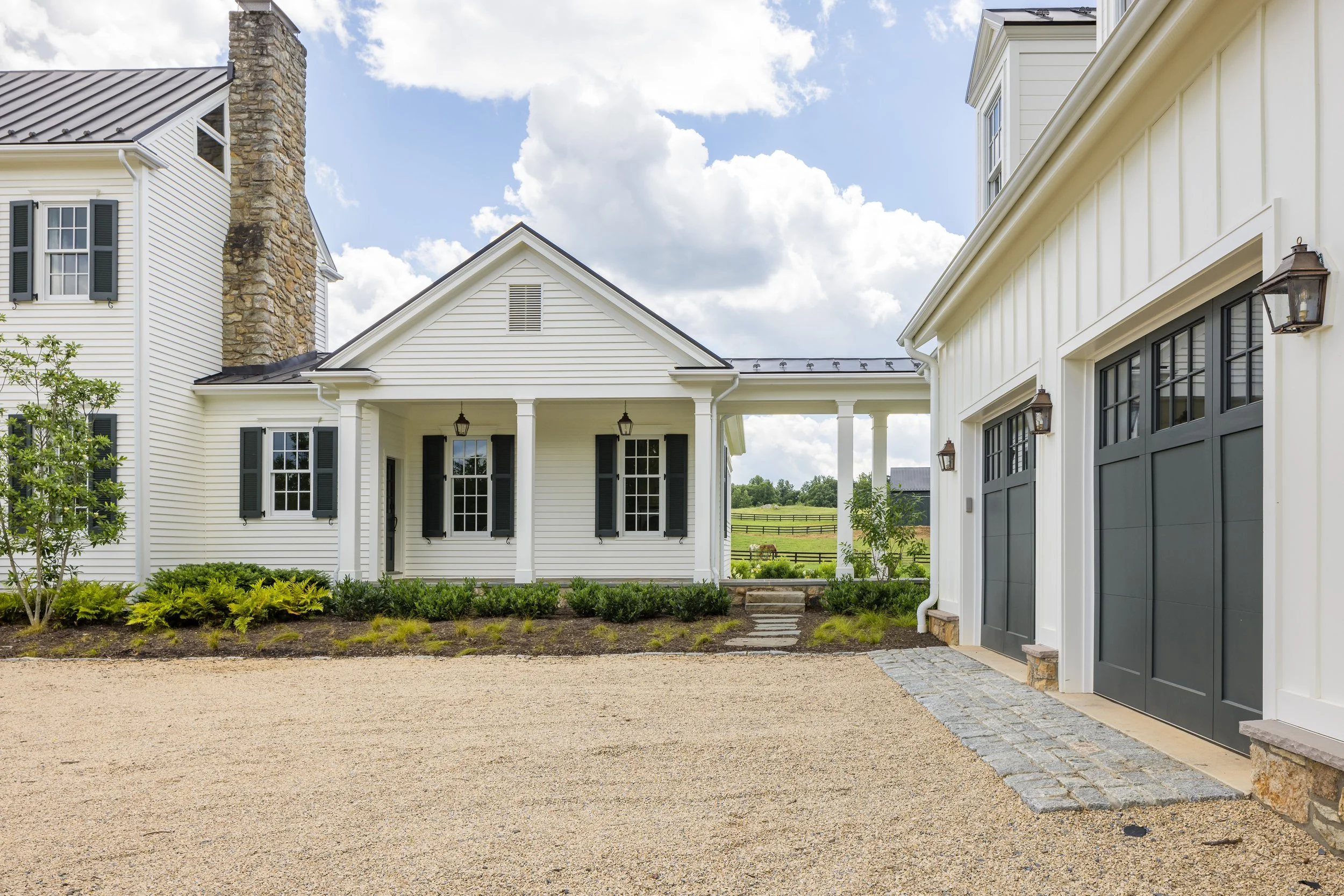 A modern farmhouse-style home with white siding, black shutters, and a black garage door, set in a landscaped yard with a gravel driveway and a view of green fields in the background.