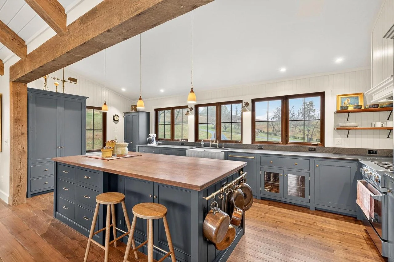 Modern kitchen with blue cabinets, wooden island, and hardwood floors. Large windows with a view of outdoors, pendant lighting, and open shelves with dishes.