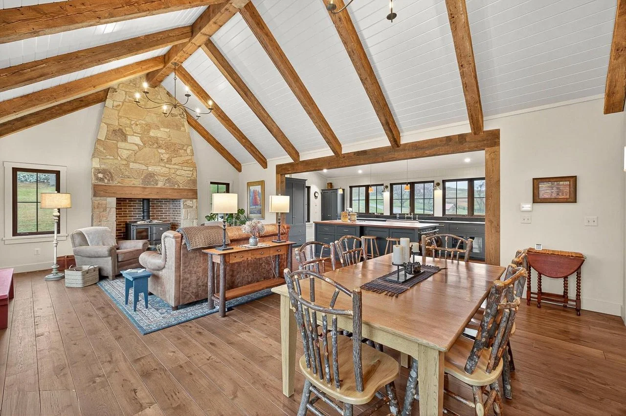 Open living and dining room with wooden beams, stone fireplace, large windows, and rustic furniture.