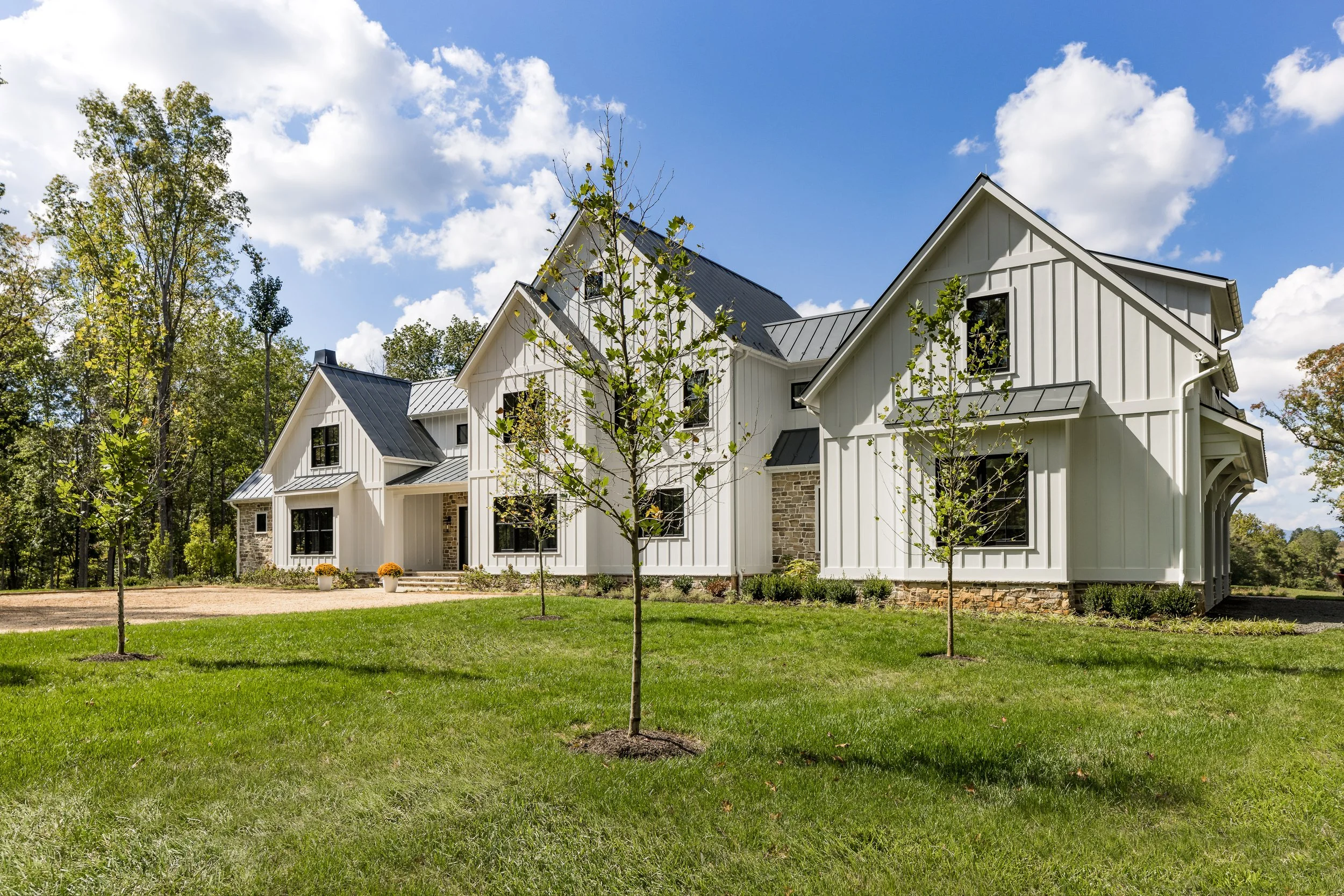 A modern farmhouse with white siding, black window frames, and a metal roof, surrounded by a green lawn and small trees, under a partly cloudy blue sky.