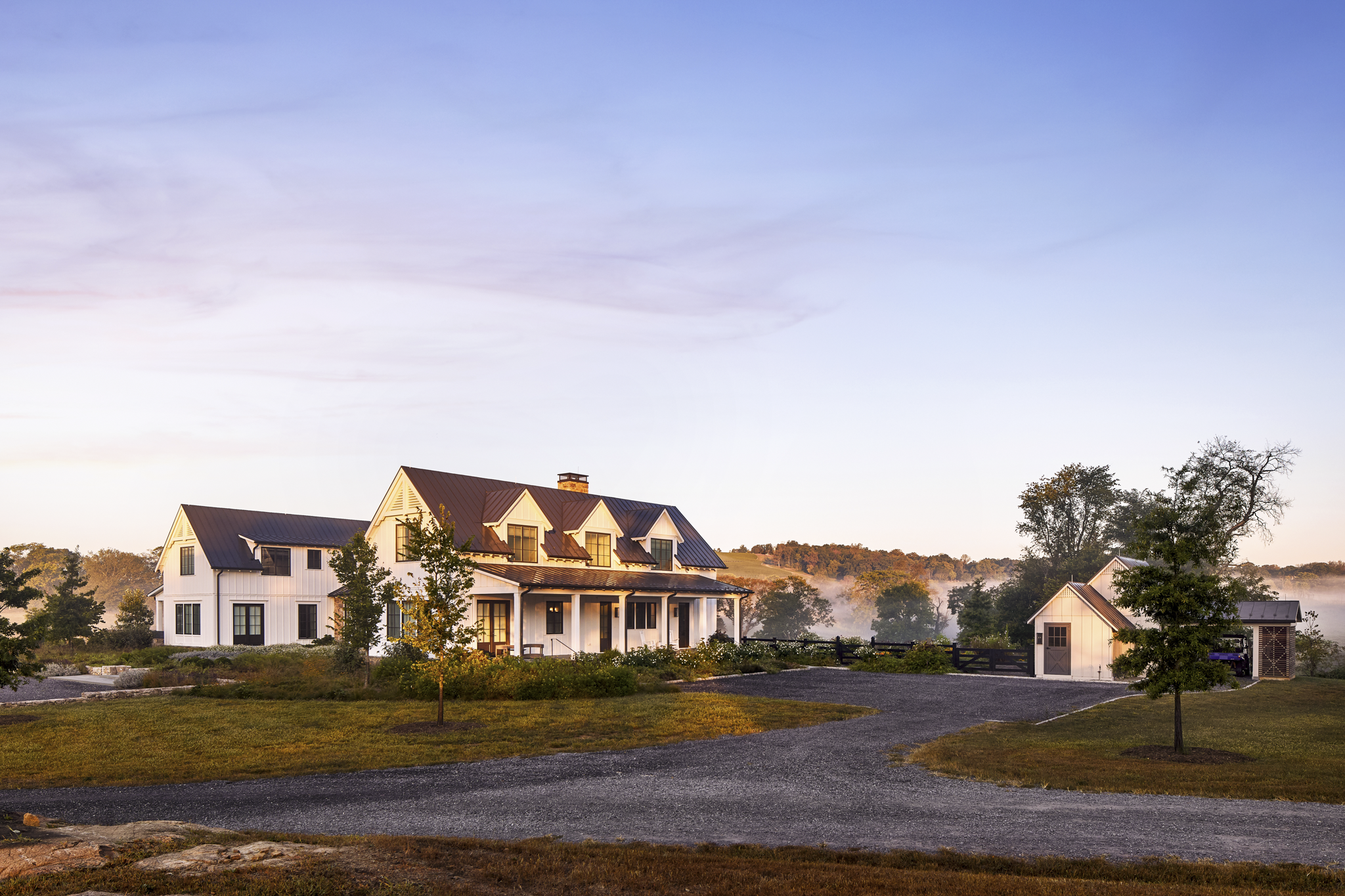 A large white house with a metal roof, surrounded by trees and a gravel driveway, with smaller outbuildings and misty hills in the background at sunrise or sunset.