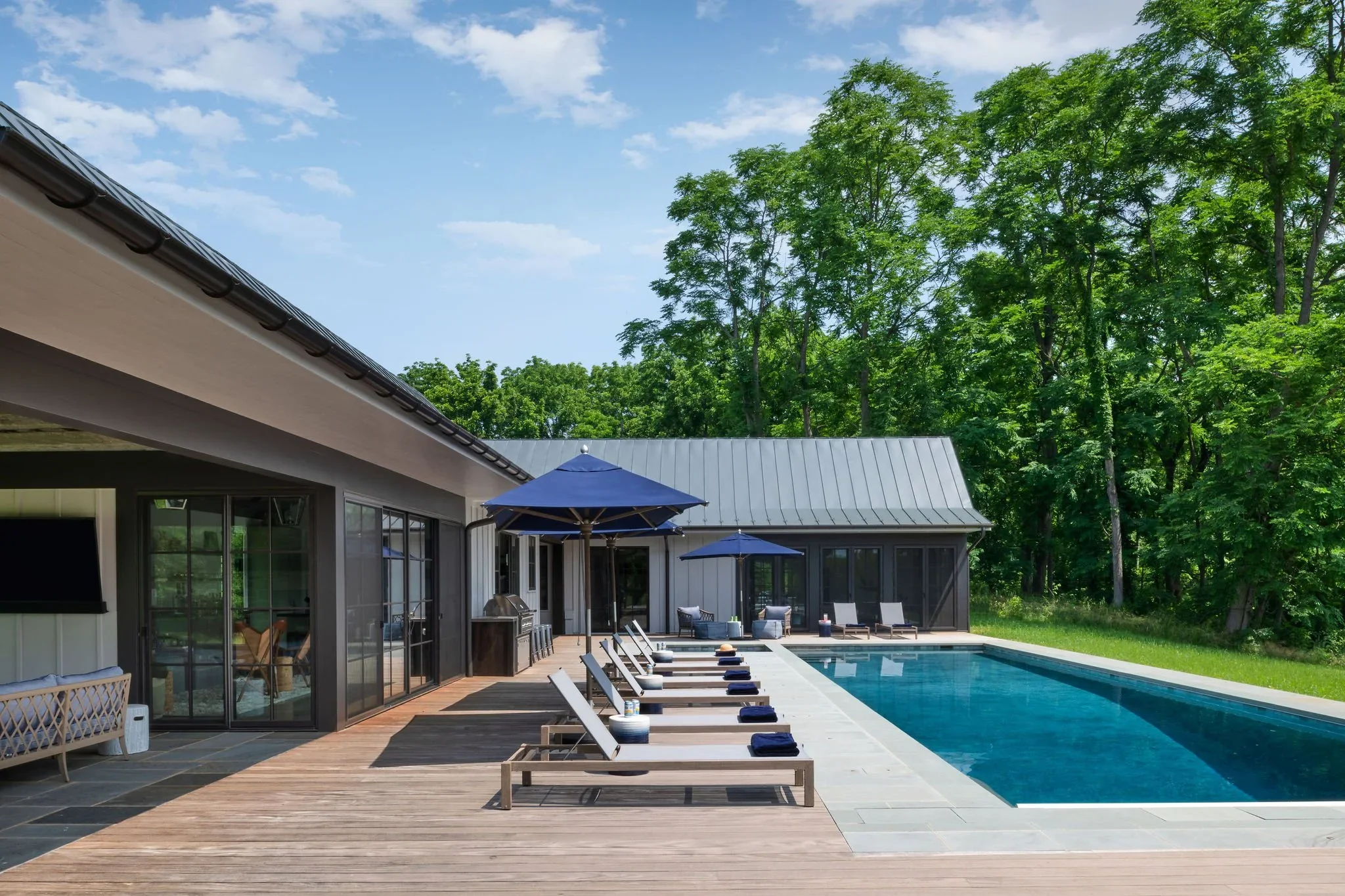 A backyard with a rectangular swimming pool, wooden deck, lounge chairs with dark towels, blue umbrellas, and a modern house with large glass doors, surrounded by green trees under a blue sky.