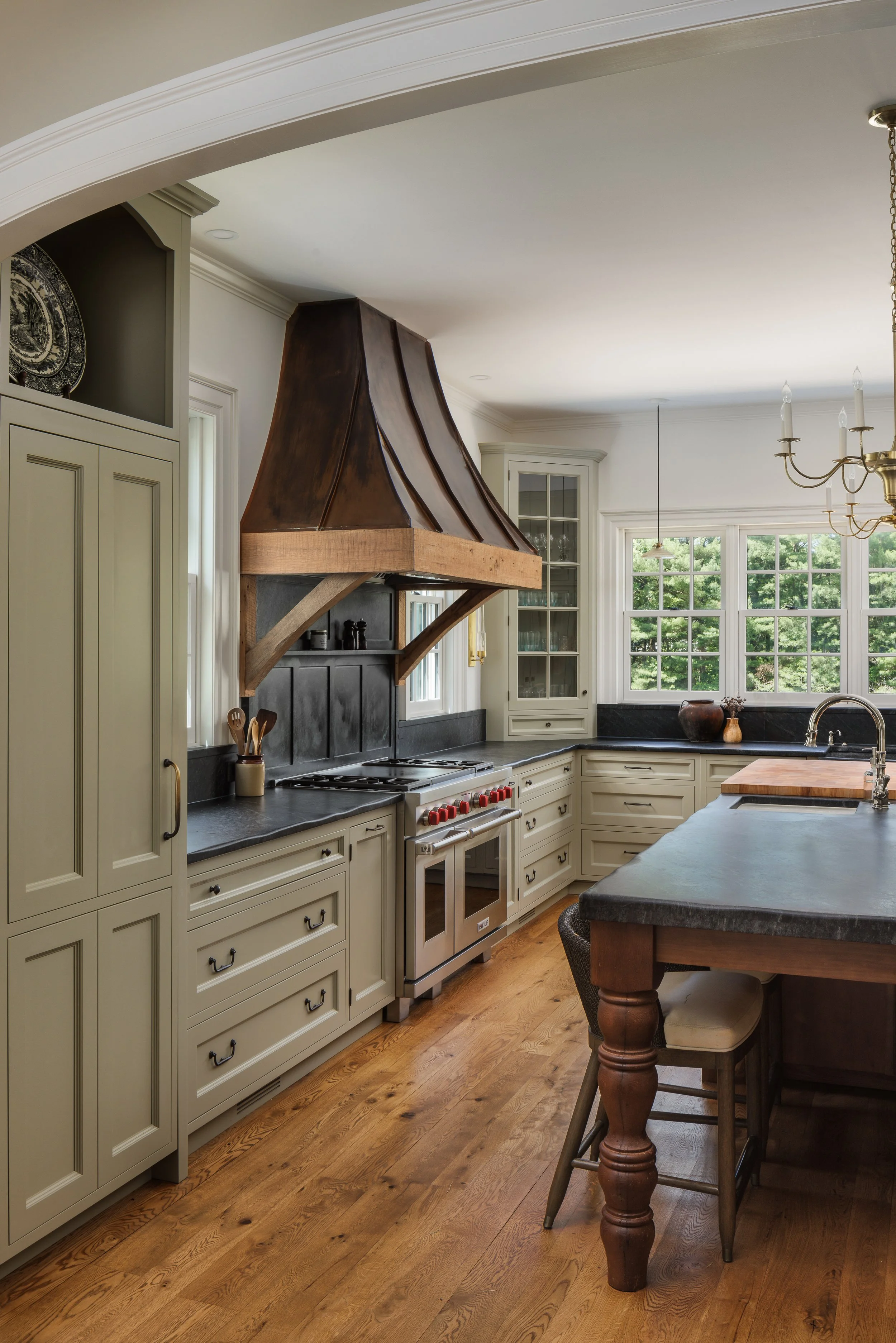 A kitchen with cream-colored cabinets, black countertops, and a stainless steel oven. A large wooden range hood is above the stove. There are windows with greenery outside, a chandelier, and a kitchen island with a wooden top.
