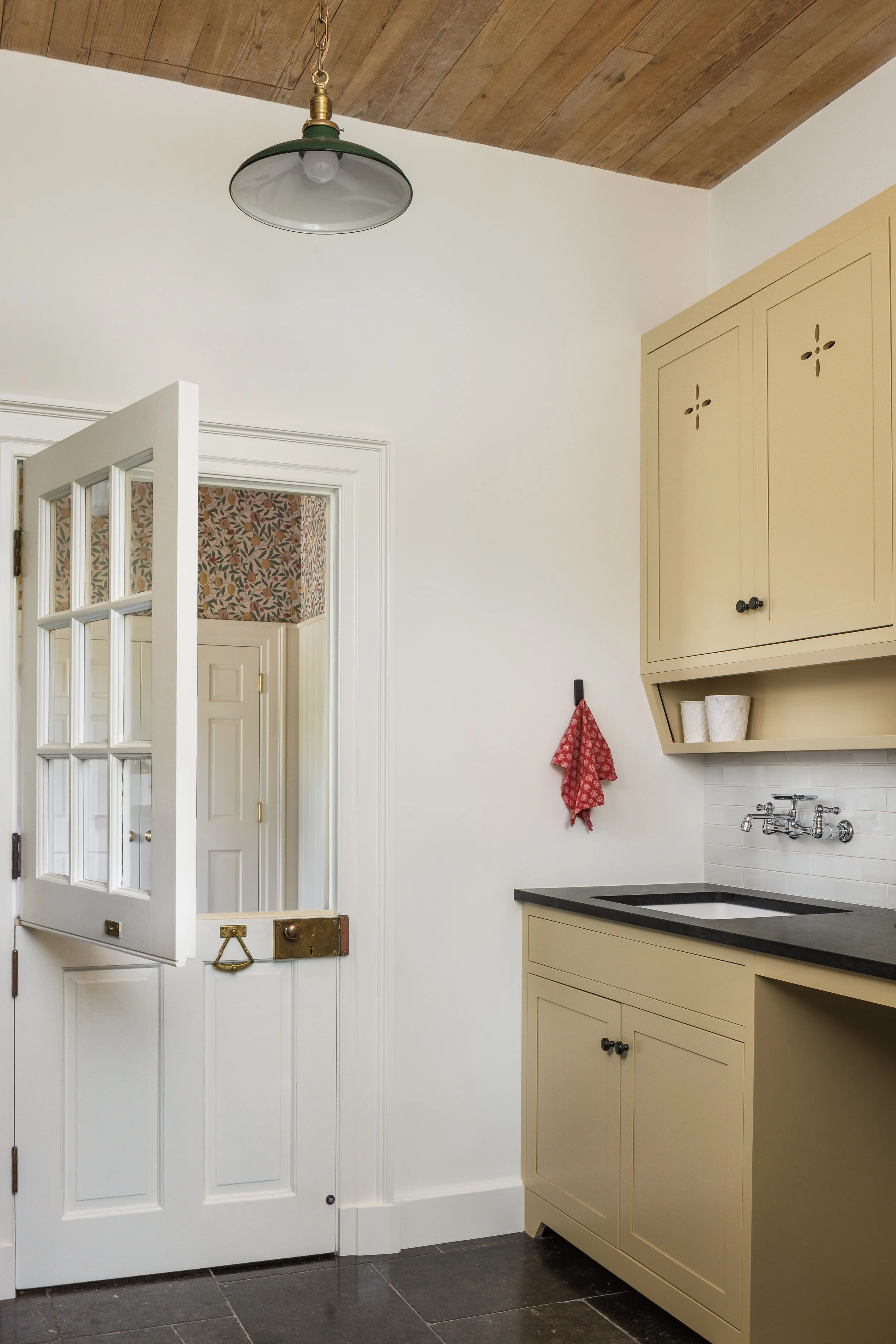 A small vintage-style laundry or utility room with cream-colored cabinets, a dark countertop, a small undermount sink, a wall-mounted faucet, a red dish towel hanging on a hook, and a ceiling light fixture. There is a partly open glass-paned door lea