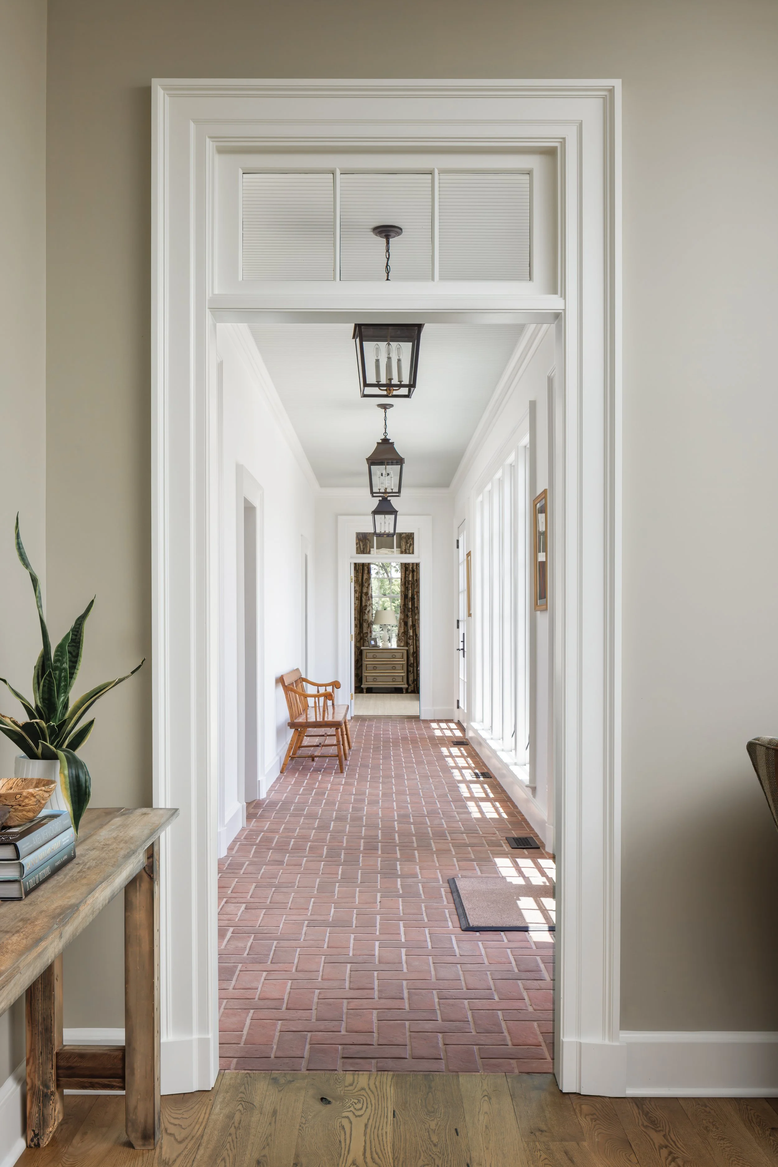 Interior view of a house hallway with brick flooring, white walls, and three black lantern-style ceiling lights. A wooden bench is positioned on the left side near archways, with curtains and a window at the end of the hallway.