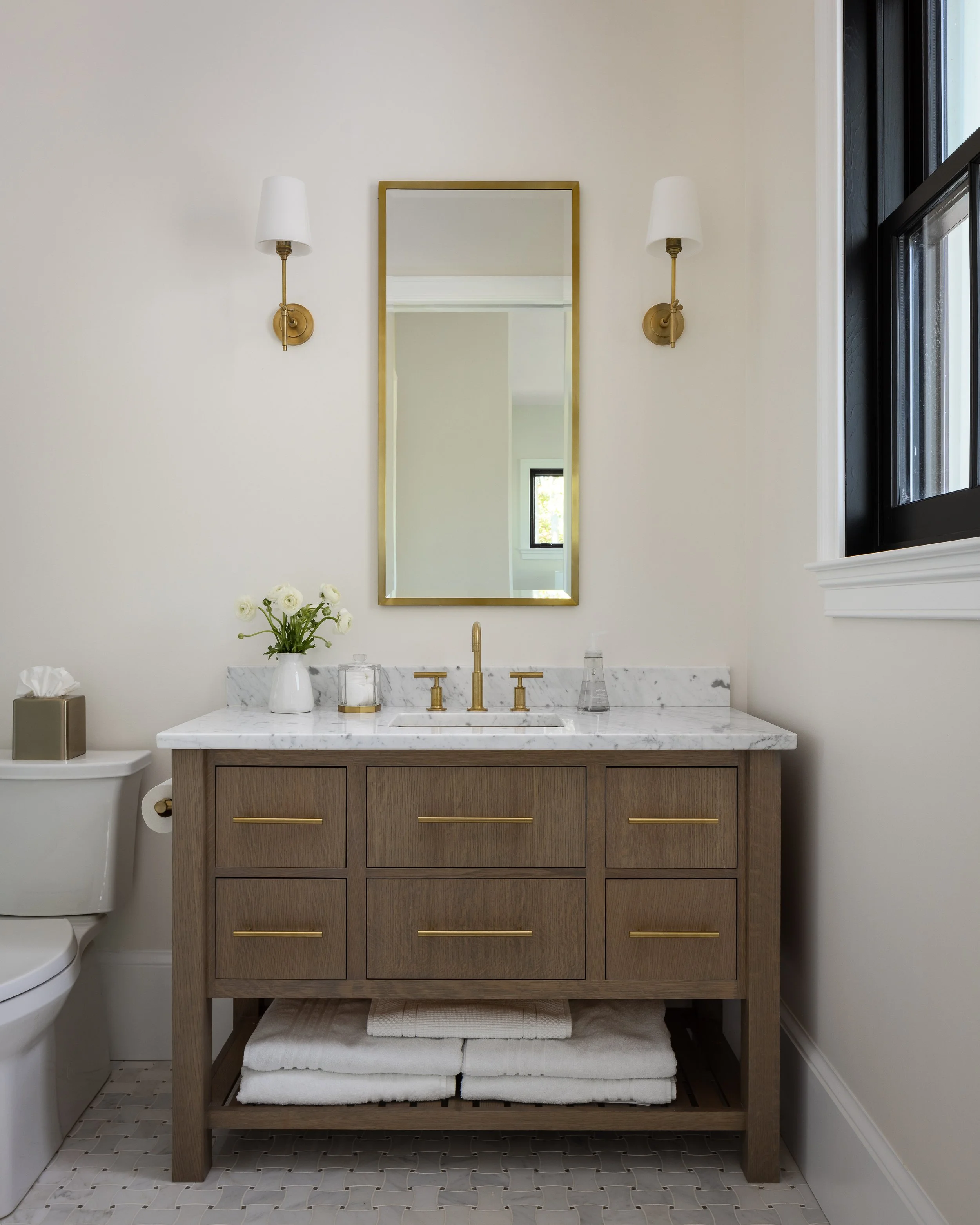 Bathroom vanity with a marble countertop, gold fixtures, a mirror, wall sconces, and towels underneath. A toilet is visible on the left, and a window is on the right.