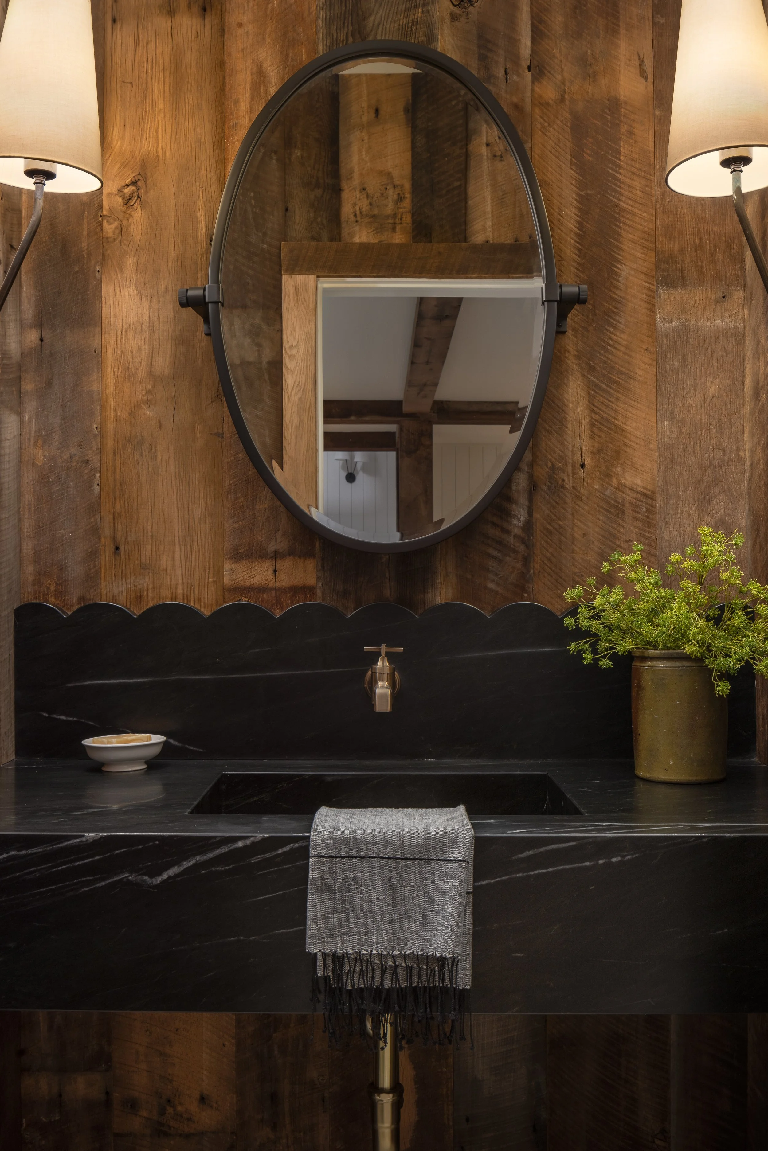 A bathroom vanity with a black marble countertop, a black oval mirror, potted green plant, and beige wall sconces.