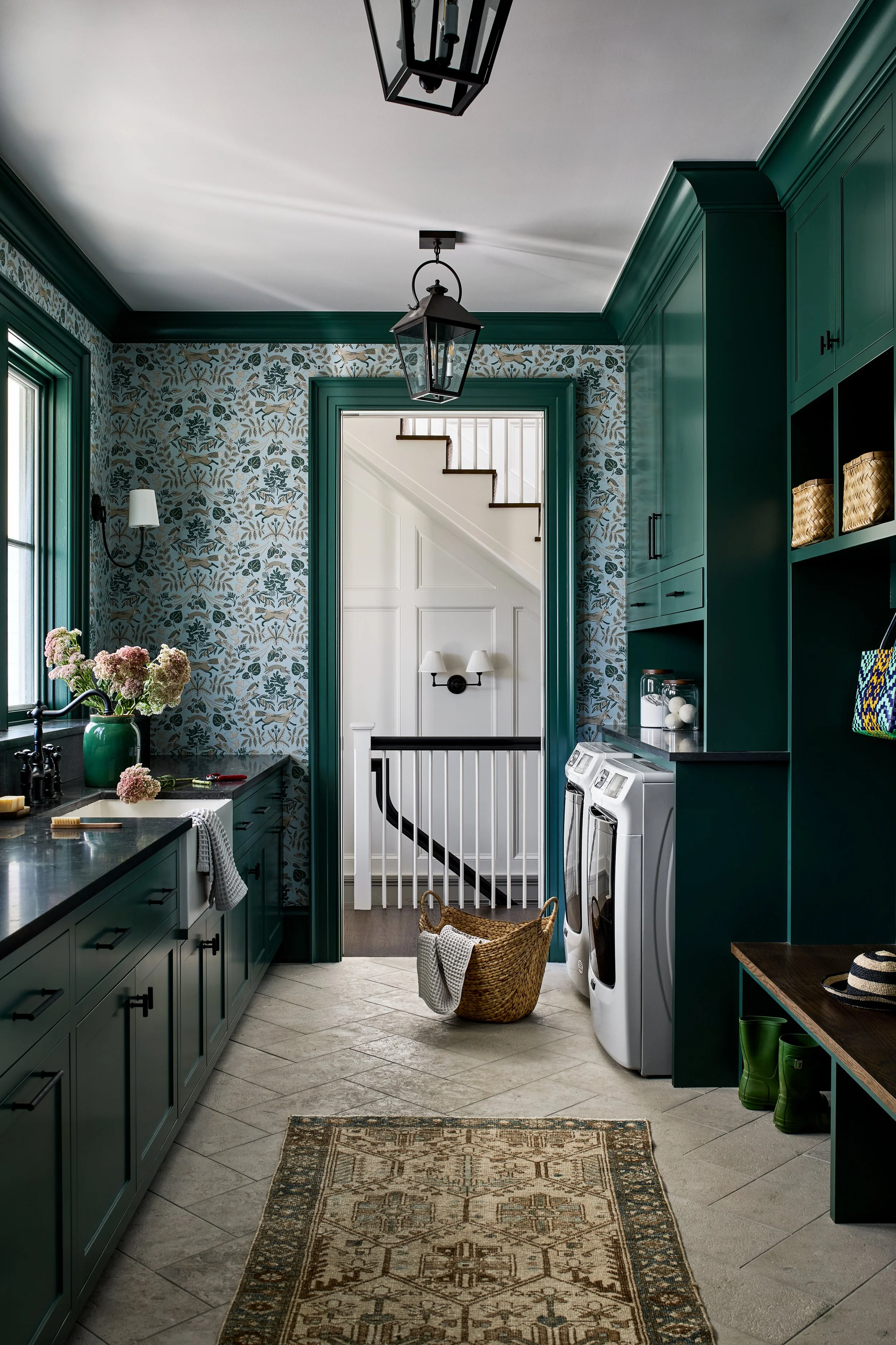 A laundry room with green cabinetry, wallpaper with floral pattern, and a white laundry machine. There is a basket with a towel, green boots, and a hat on a bench. A window with vase of flowers, pendant lights, and a staircase visible through a doorw
