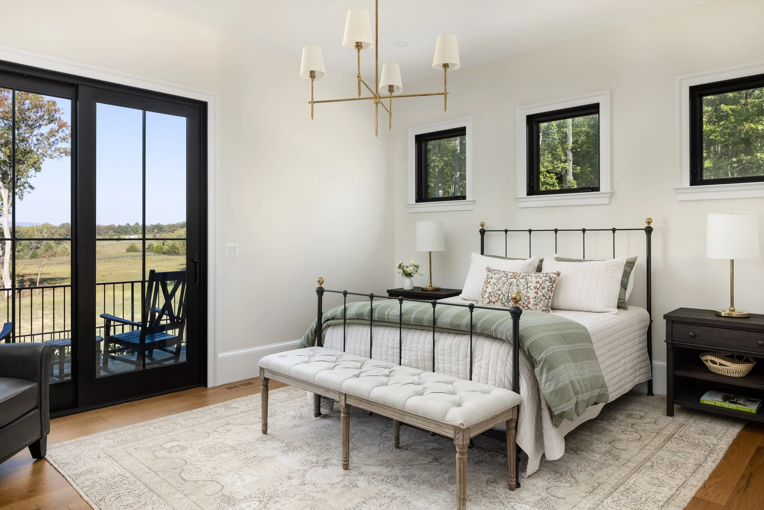 A bedroom with a black metal bed, white and patterned pillows, a bedside table with a lamp, a bench at the foot of the bed, and three small windows on the wall. There are two lamps on the bedside tables, one on each side. A sliding glass door leads t