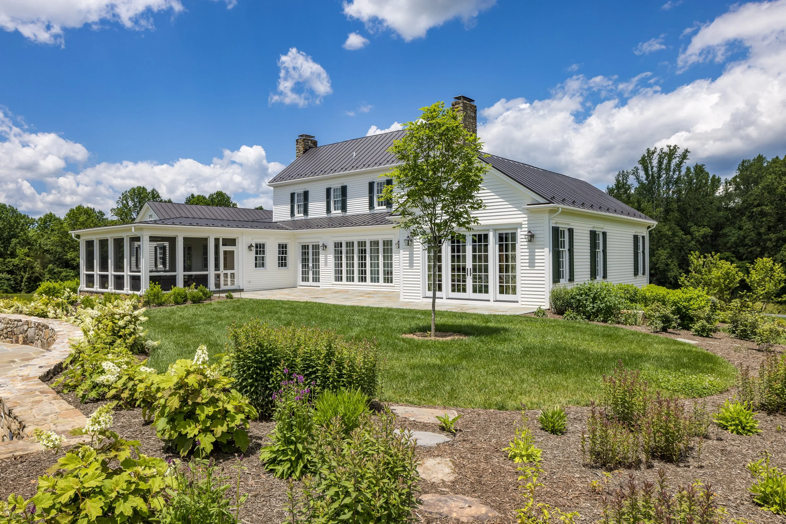 A large white house with black shutters and a dark gray roof, surrounded by a well-maintained garden with various plants and a small tree, under a partly cloudy blue sky.