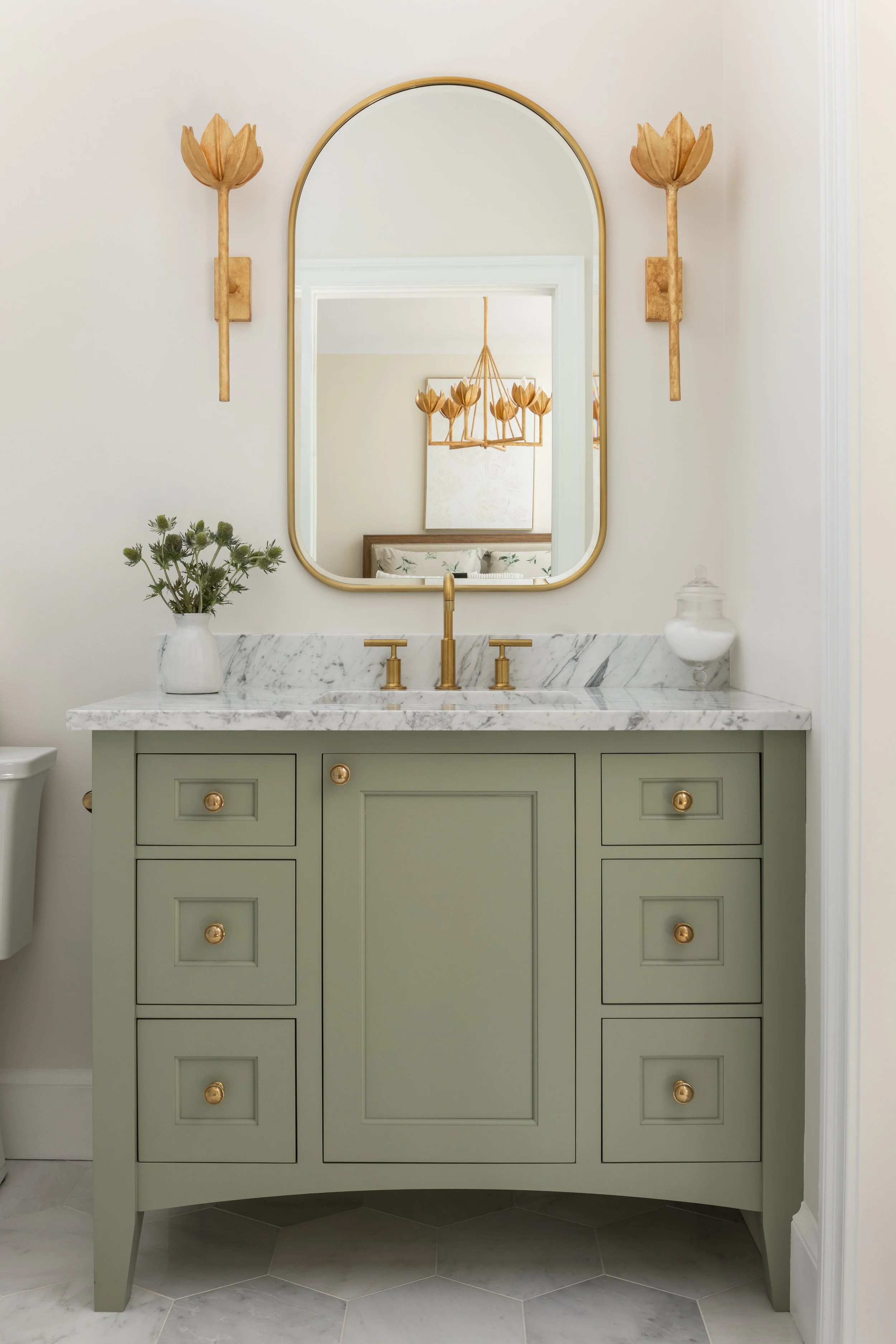 A bathroom vanity with light green cabinets, a white marble countertop, and gold hardware. A gold faucet is centered under a gold-framed mirror, with wooden flower wall sconces on either side. There is a small plant in a white vase and a glass contai