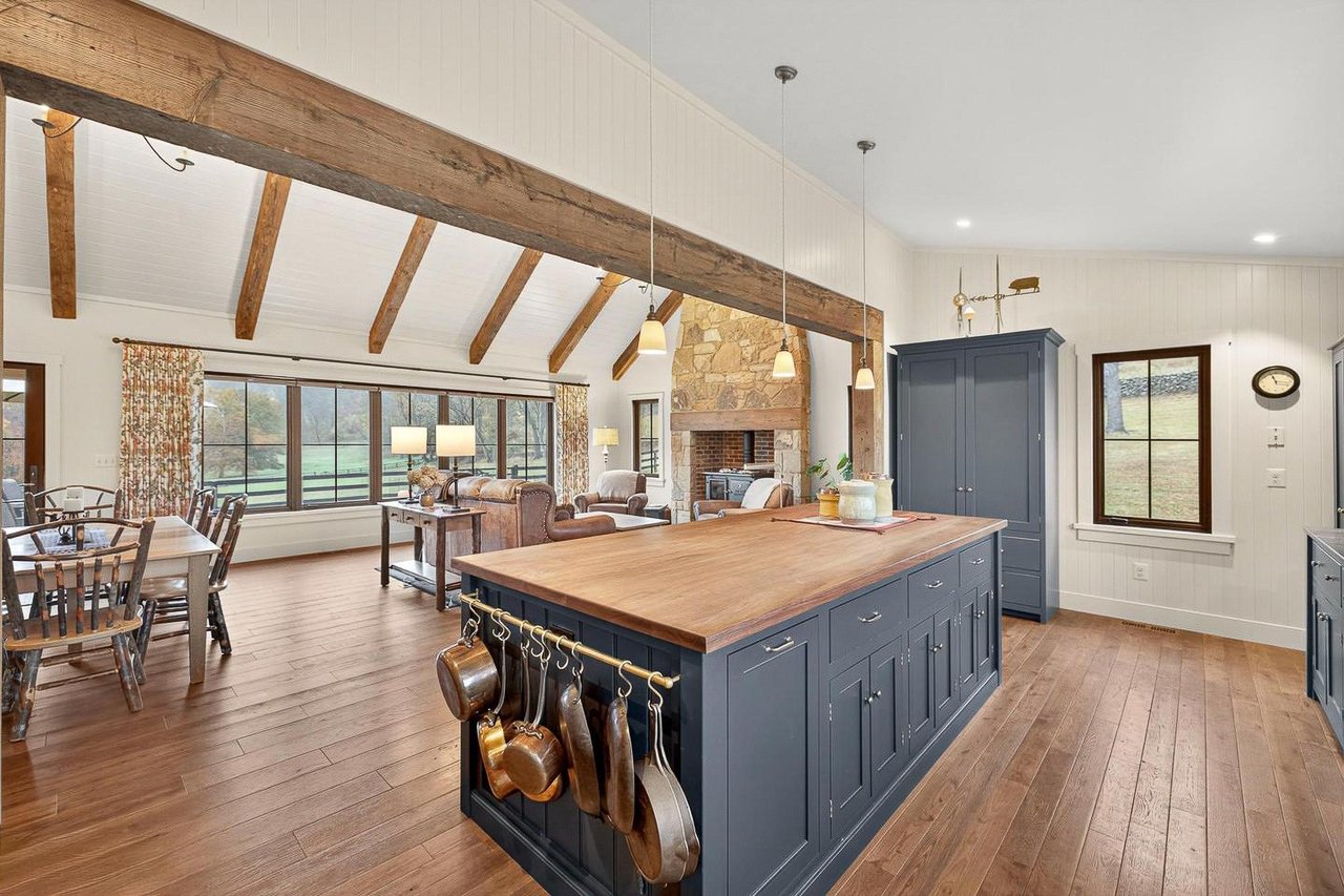 Open-concept kitchen and living room with wooden floors, a blue kitchen island, a stone fireplace, large windows, and dining and living areas with furniture and decor.