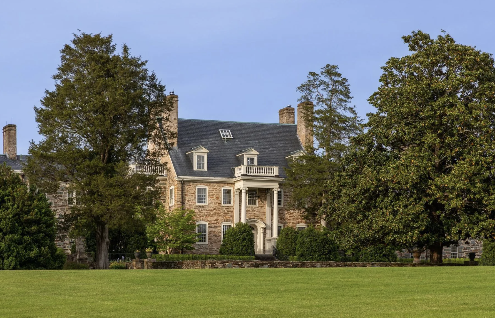 A large, historic stone mansion with a slate roof, surrounded by lush trees and a well-manicured lawn.