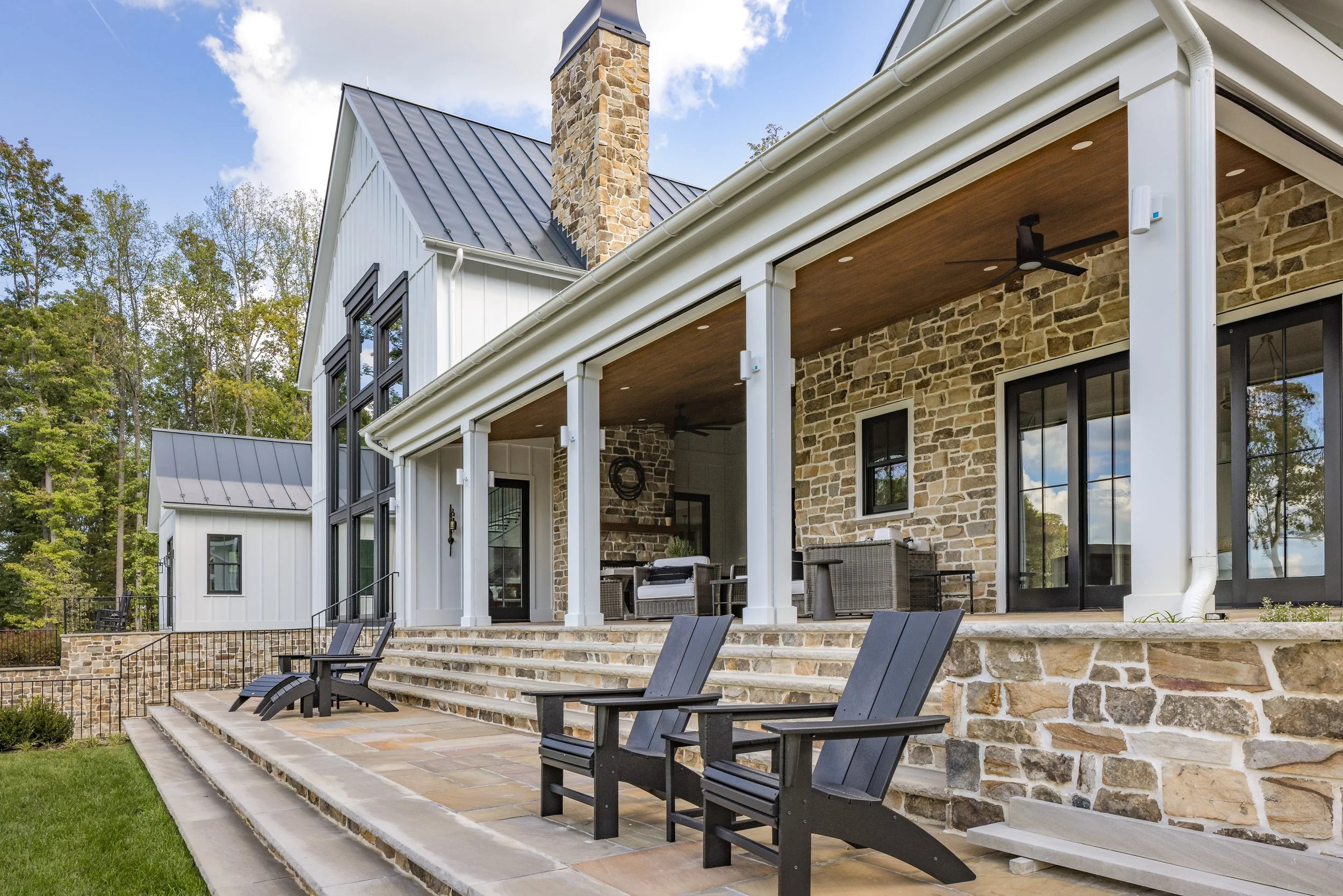 A modern house with a stone and white exterior, black framed windows, a covered porch with ceiling fans, and outdoor seating on a stone-paved patio surrounded by lush green trees.