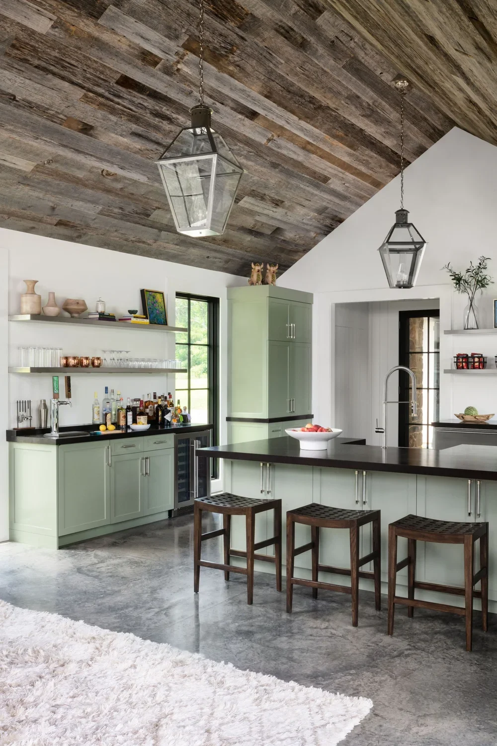 Modern kitchen with green cabinets, black countertop, wooden stools, and two pendant lights hanging from a wooden ceiling.