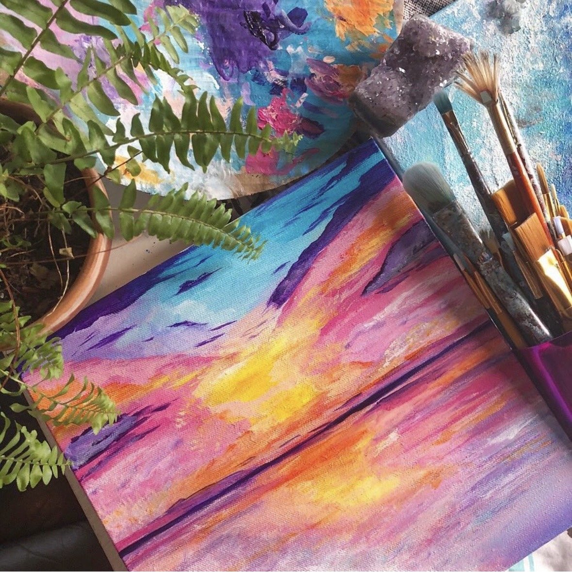 A colorful painting of a sunset over the ocean sits surrounded by paintbrushes, a fern and a crystal, by artist, Dana Savoca Tietze