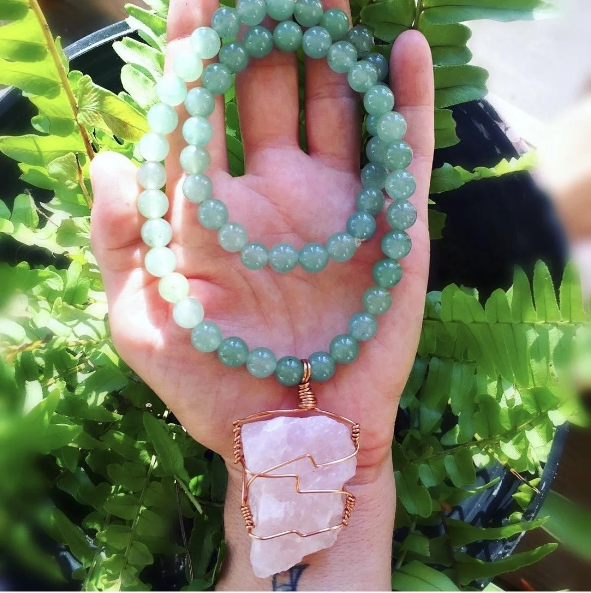 A beaded necklace with a rose quartz pendant held in an open palm above a fern