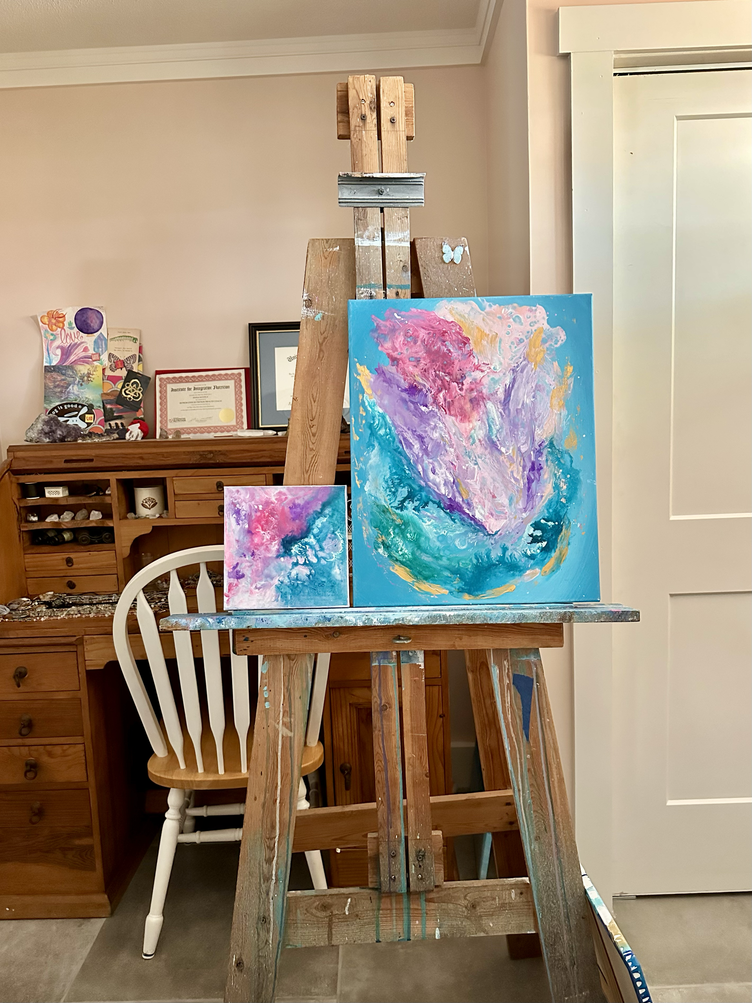 Two coordinating pink and turquoise paintings sit on a wooden easel inside an artist studio