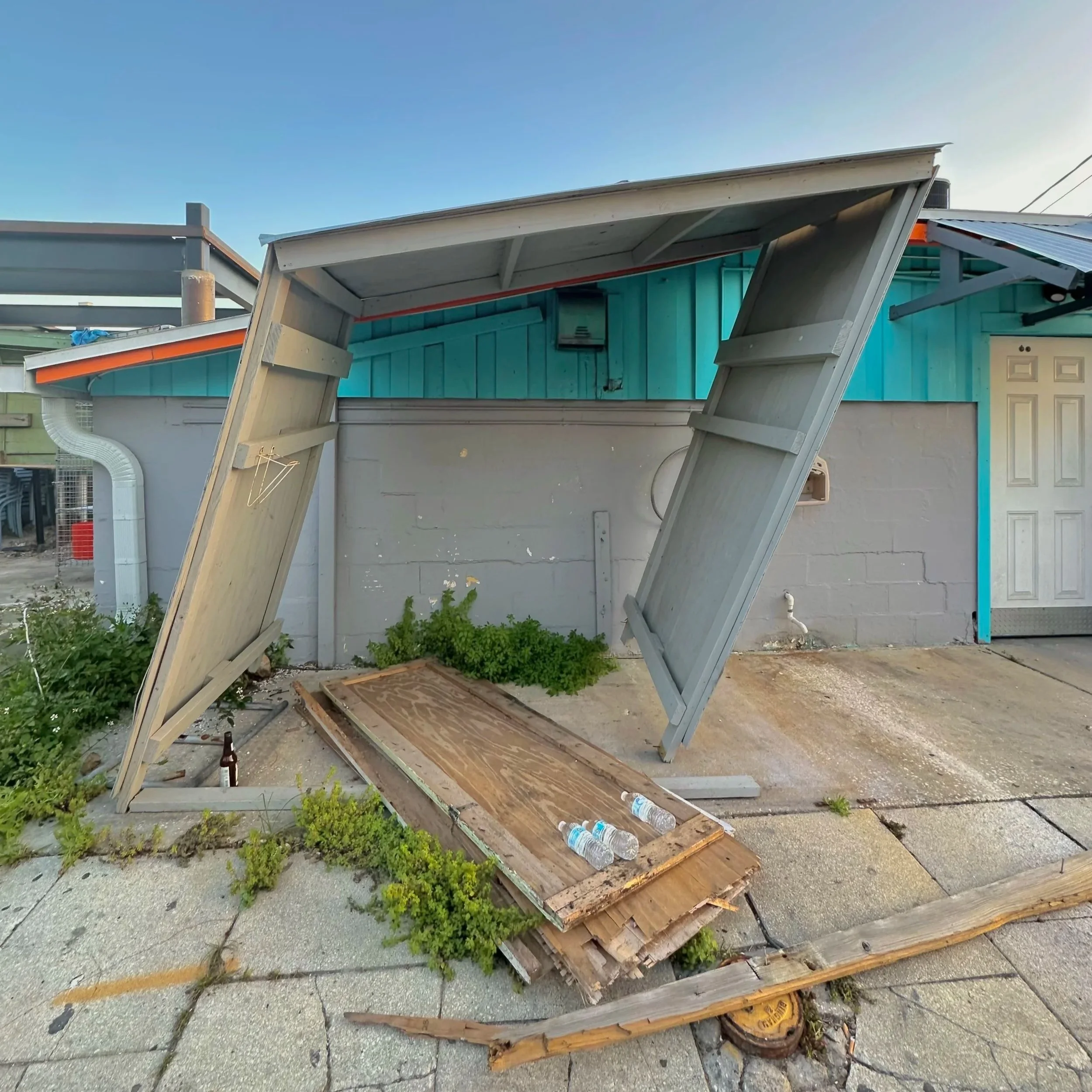 A crooked and broken structure leans dangerously behind a building in an alley way littered with plastic water bottles and beer bottles