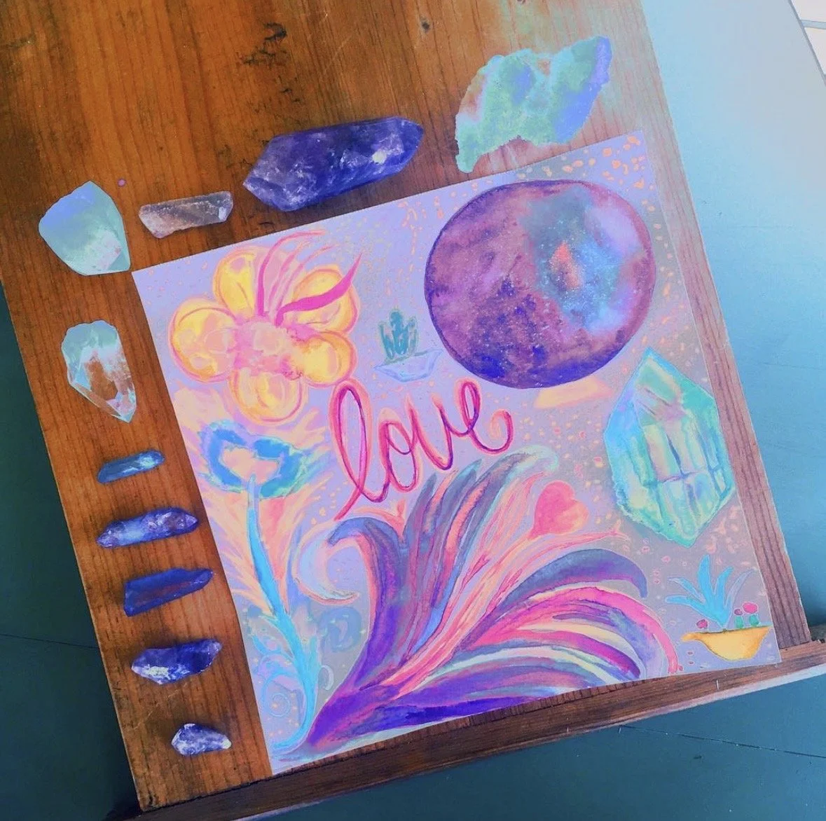 A Dana Savoca Tietze original floral watercolor is displayed on wood surrounded by crystals