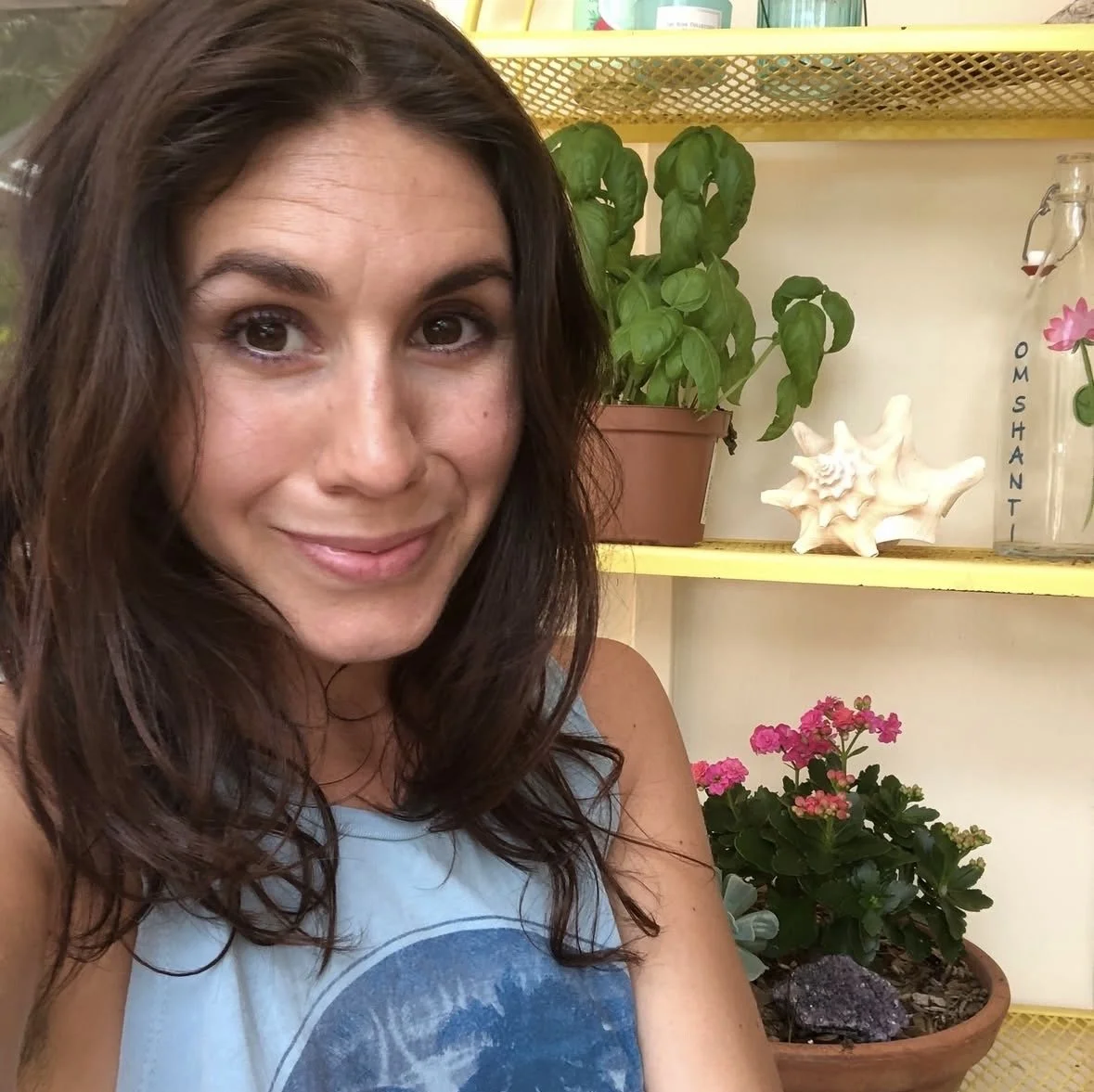 A woman with dark brown wavy hair smiling in front of a yellow shelf with potted plants, a white sea shell sculpture, and a pink flowering plant.