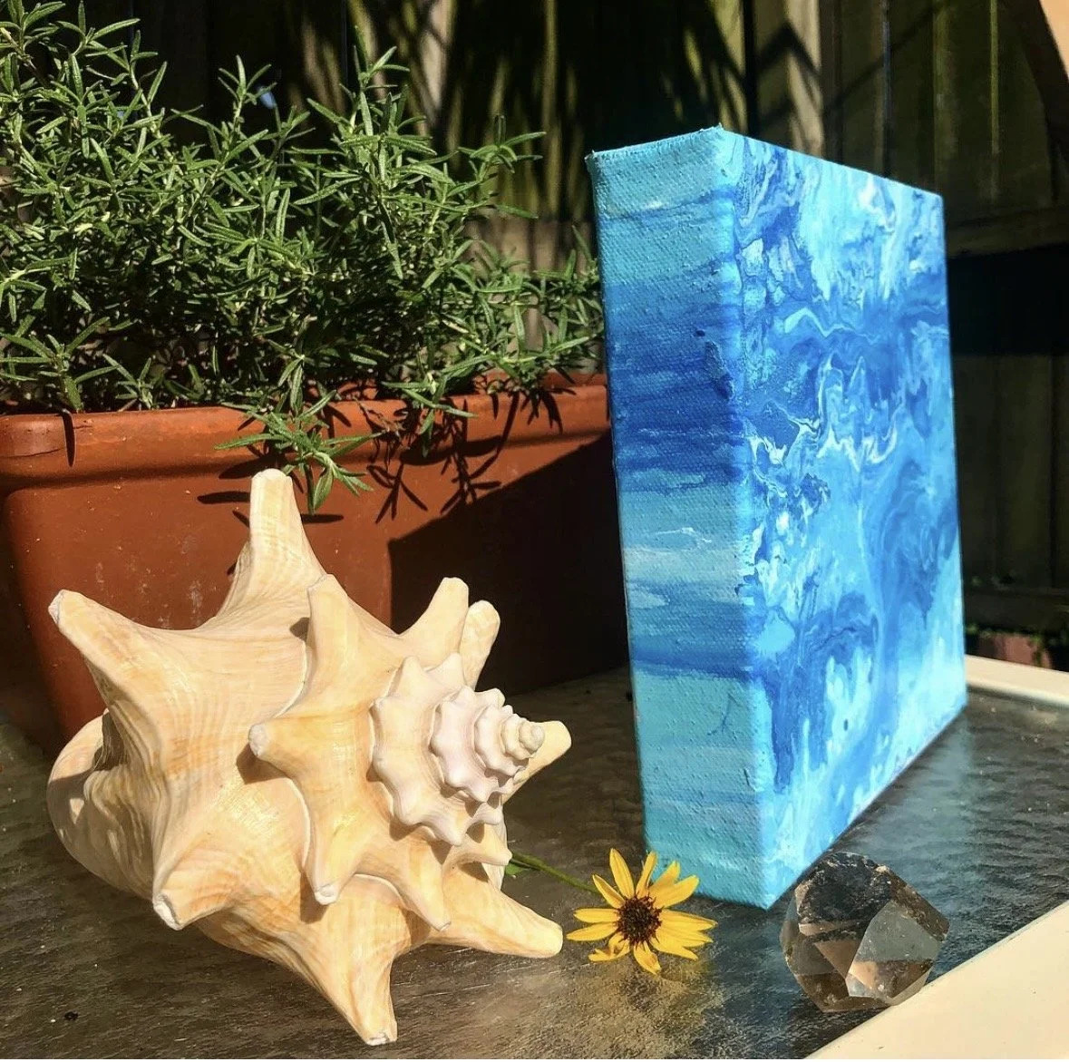 An abstract fluid art canvas in oceanic blue tones sits in a garden with flowers and a large shell