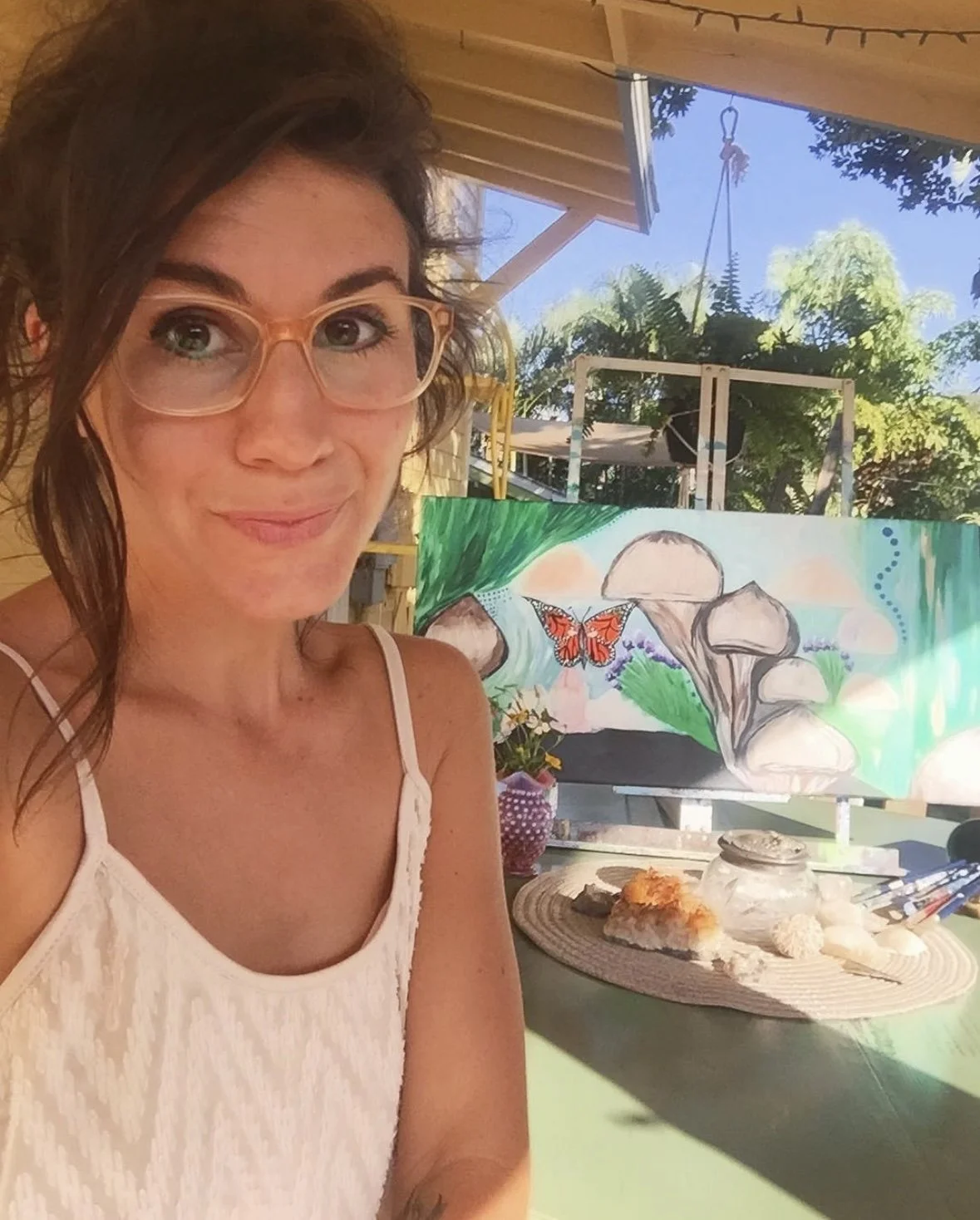 Artist Dana Savoca Tietze sits outside in front of a painting she's working on that features psychedelic mushrooms, crystals and a butterfly.