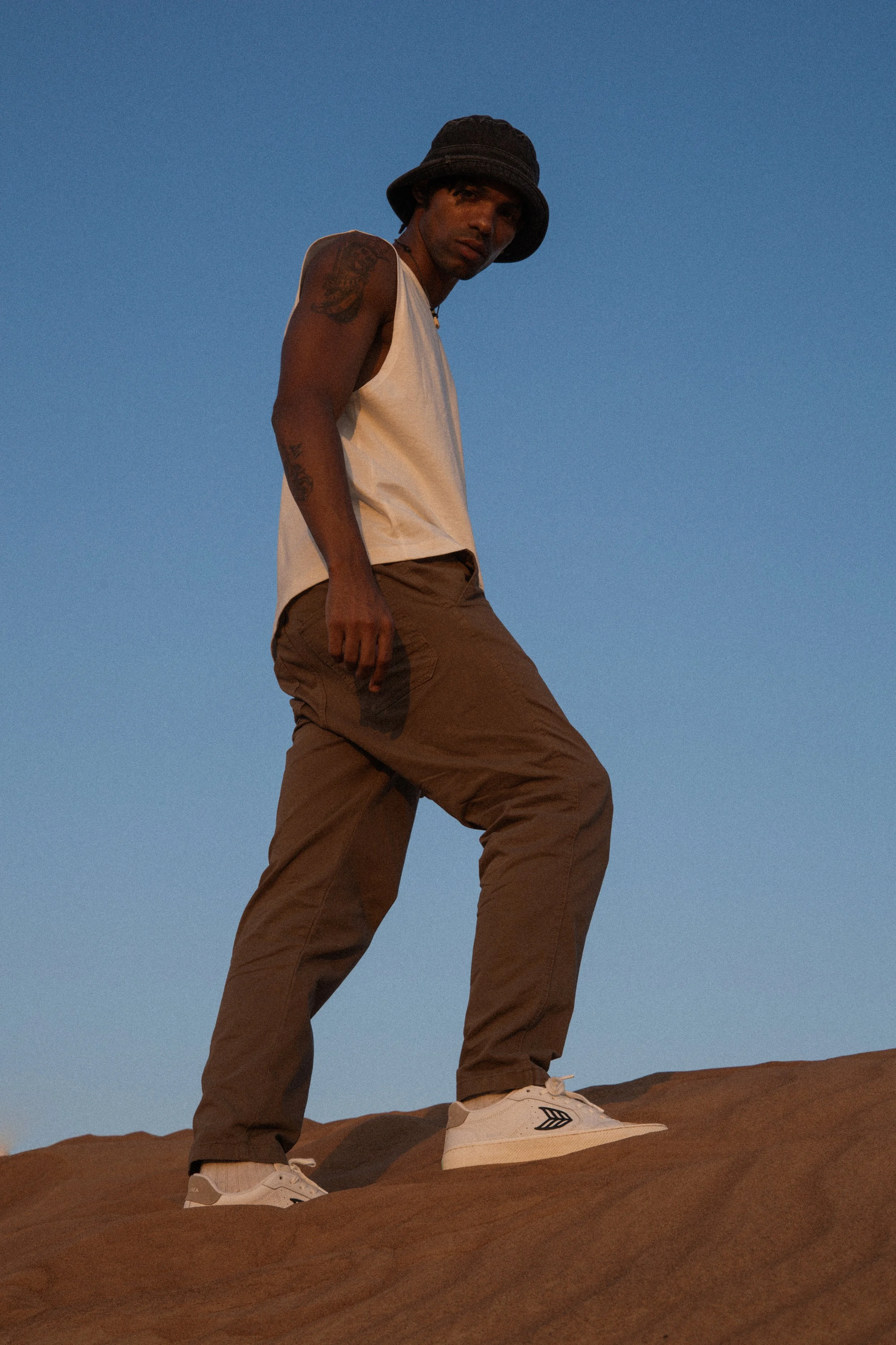 A young man wearing a black bucket hat, white tank top, brown pants, and white sneakers stands on a reddish-brown rocky surface against a clear blue sky.
