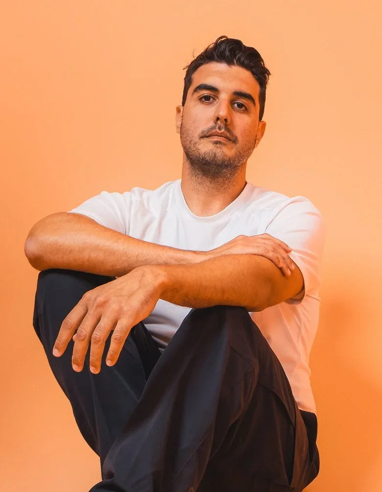 Paris Vorgias - A young man with dark hair, wearing a white t-shirt, sitting with one knee raised and resting his arm on it, against a peach-colored background.
