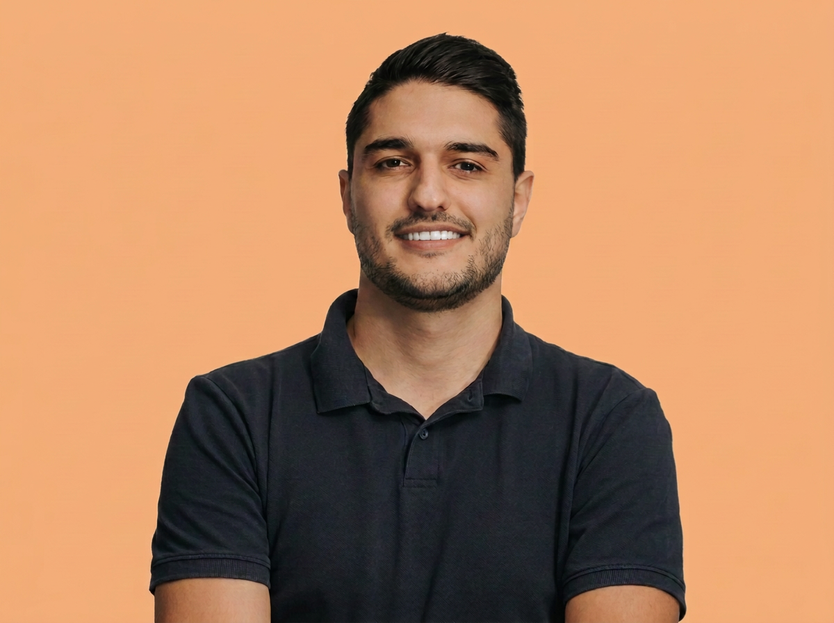 Dionisis Vorgias - A young man with dark hair, a beard, and mustache, smiling, wearing a black polo shirt, standing against a peach-colored background.