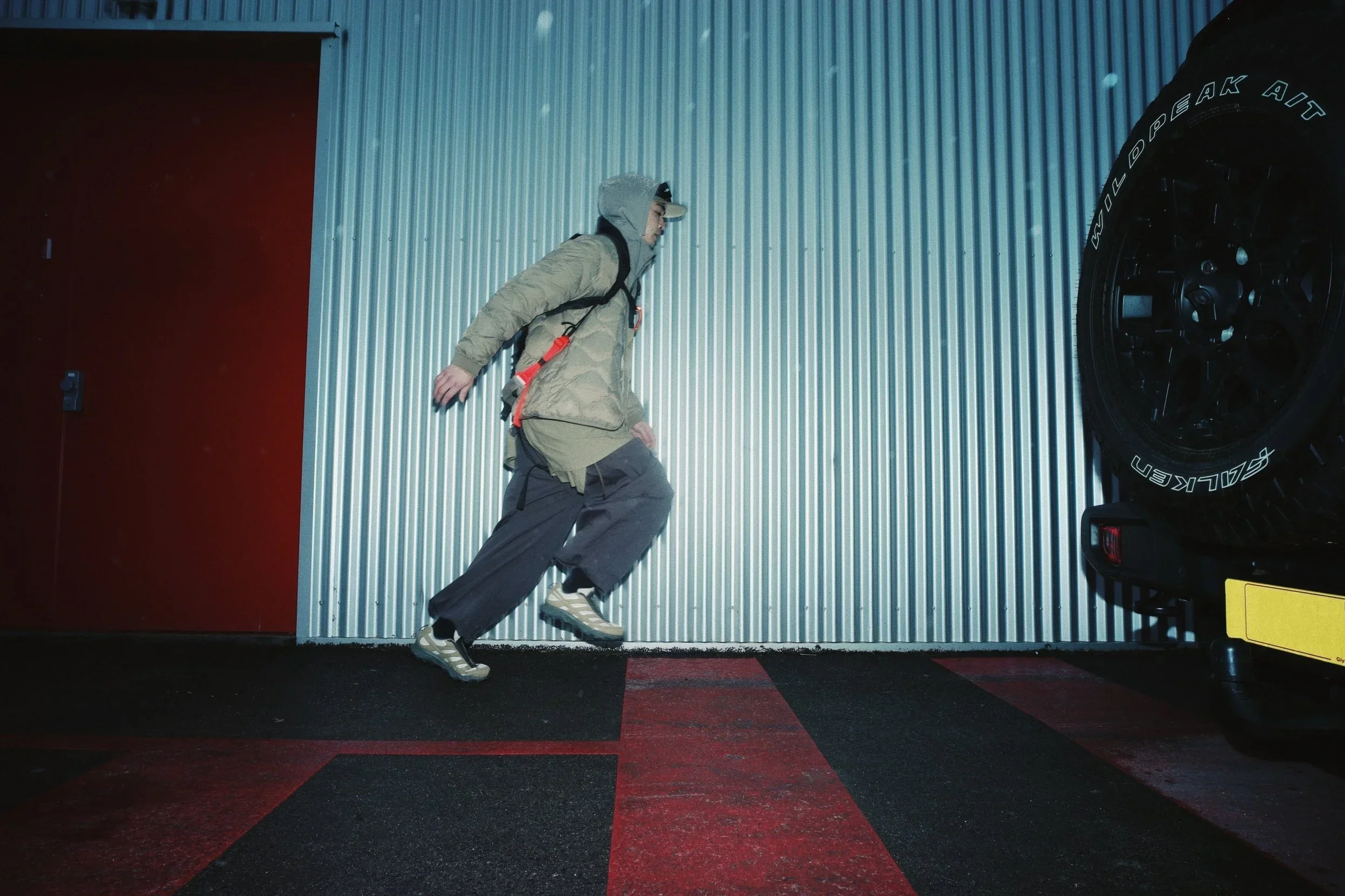 A person dressed in outdoor gear, including a tan jacket, gray pants, and hiking boots, is walking along a corrugated metal wall with a tire partially visible on the right. The scene appears to be in an industrial or garage setting with a red door on the left and a black and red carpeted floor.