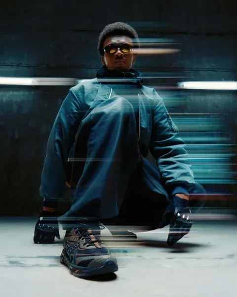Person wearing sunglasses, blue jacket, and sneakers, crouching in an urban setting with light streaks in motion blur, creating a dynamic effect.