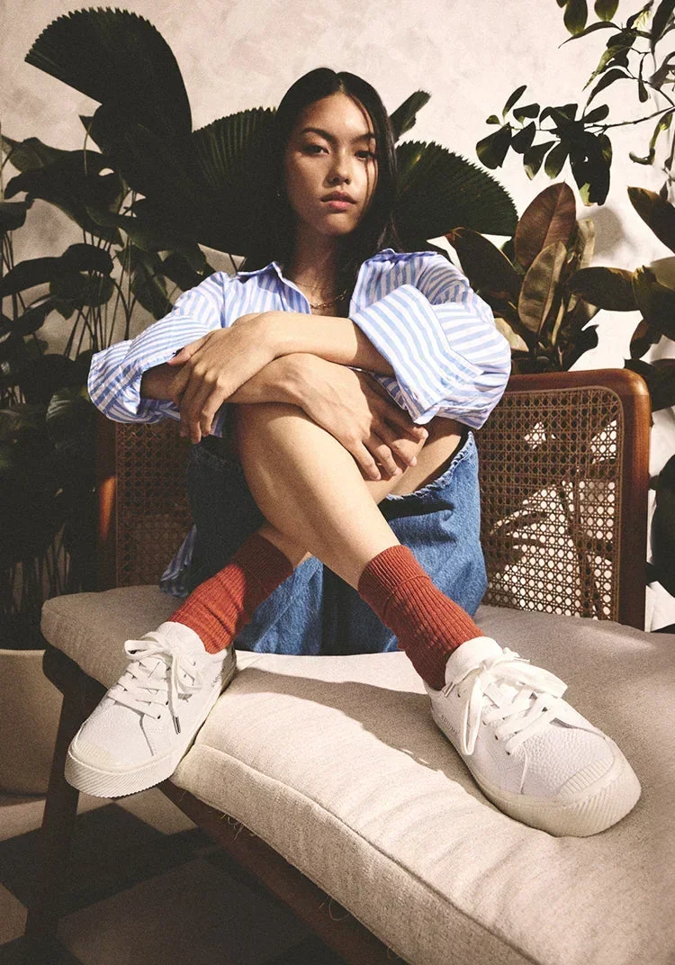 A young woman with long dark hair is sitting on a beige cushion on a wooden bench. She is wearing a blue and white striped shirt, denim shorts, orange socks, and white sneakers. She is sitting with her legs crossed and arms wrapped around her knees, looking serious. There are large leafy green plants behind her.