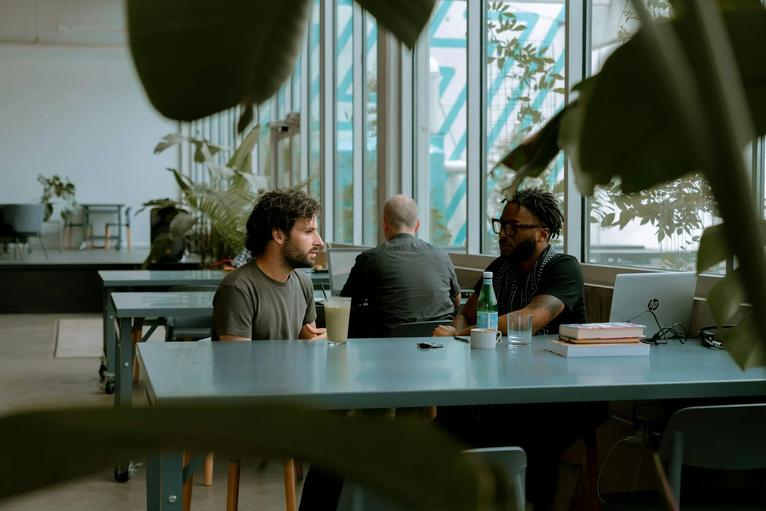 Two men sitting across from each other at a table in a bright, modern cafe with large windows and plants in the background, engaged in conversation.