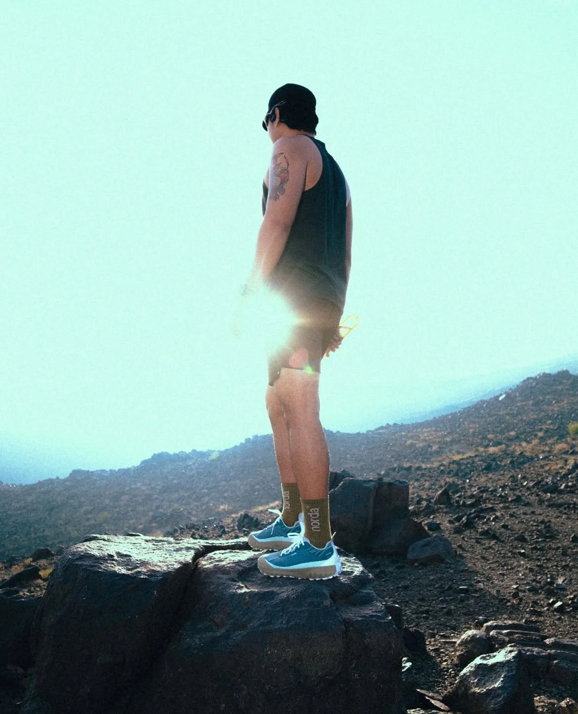 A person standing on a rock in a rugged outdoor setting, facing away from the camera, wearing sunglasses, a sleeveless top, shorts, and colorful sneakers, with the sun shining brightly behind them.