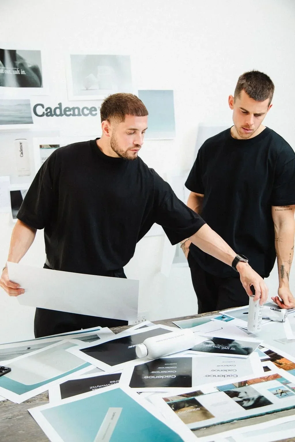 Two men in black shirts working together on a project in a bright room with a white wall, discussing design materials spread out on a table.