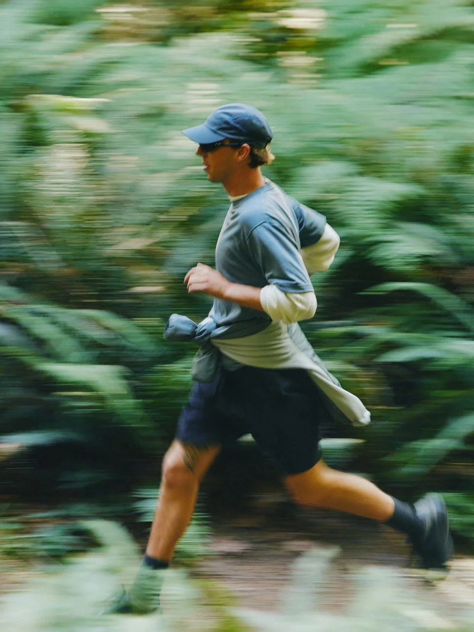 A man running through a forest trail, wearing a blue cap, sunglasses, a gray T-shirt, navy shorts, and running shoes, with a jacket tied around his waist.