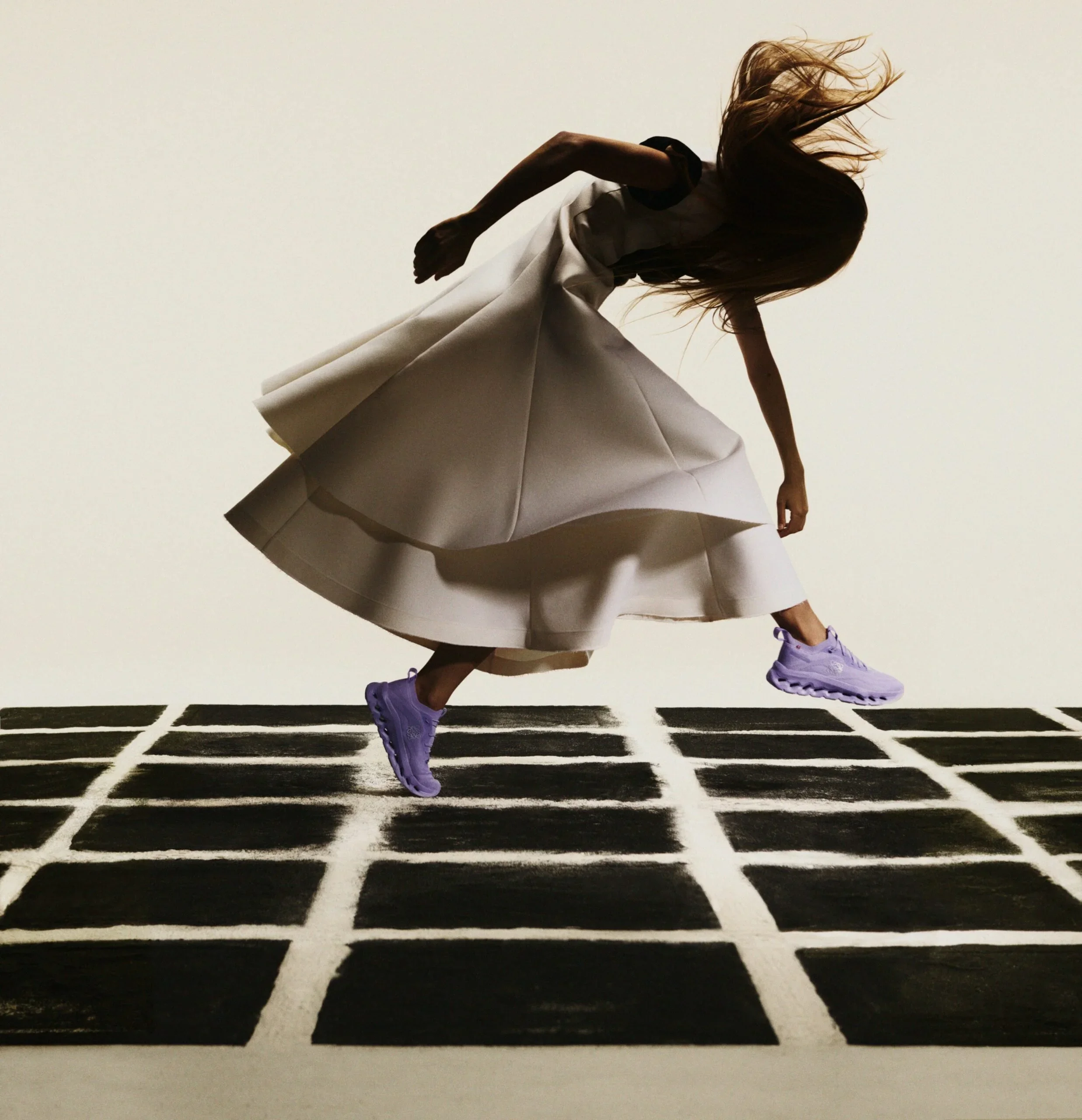 A person in a white and black outfit running or jumping on a black and white striped surface, wearing purple sneakers and with long hair flying behind.
