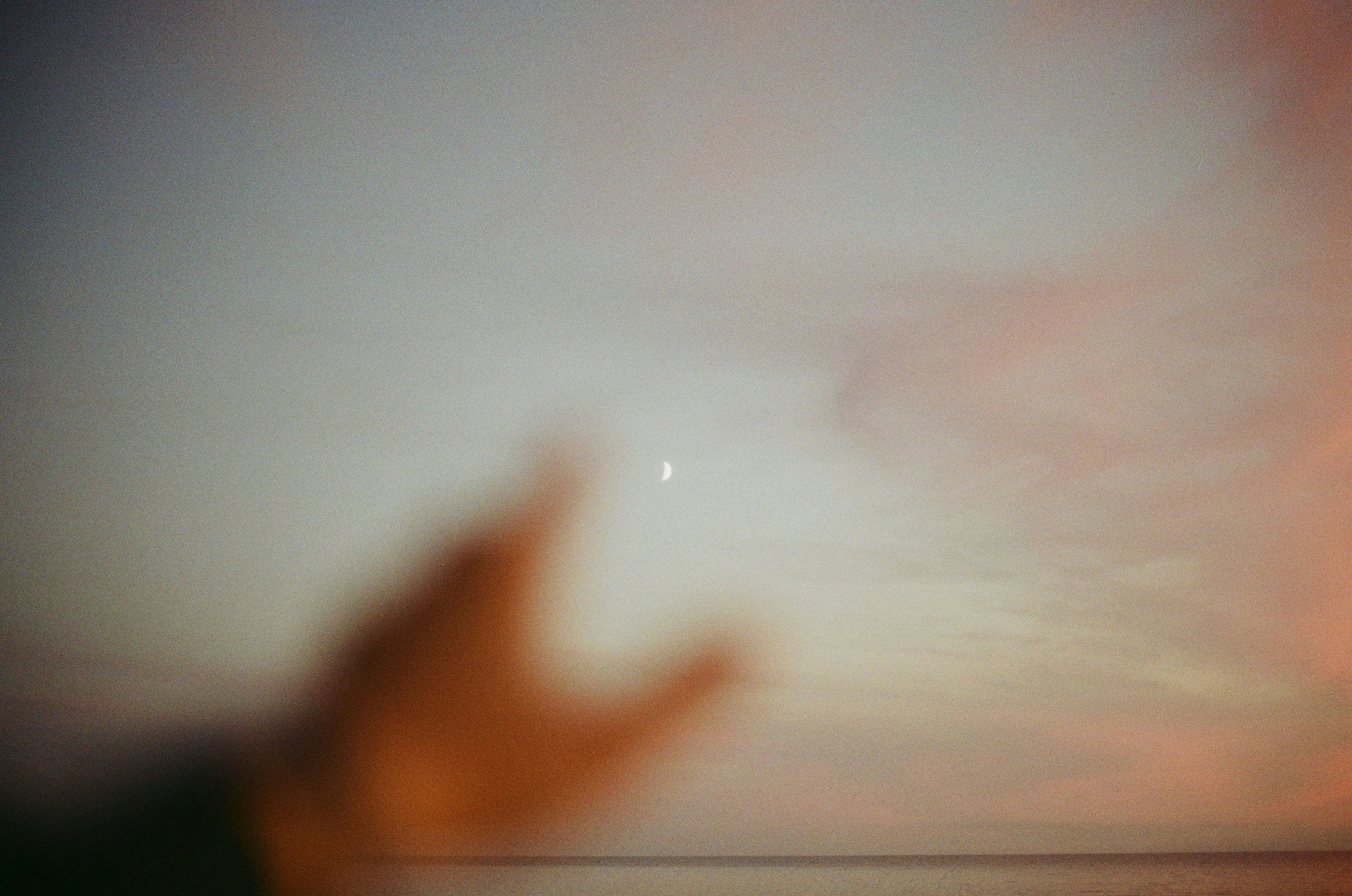 A blurred hand reaching towards the sky with a crescent moon visible in the background, during sunset or dusk over a body of water.
