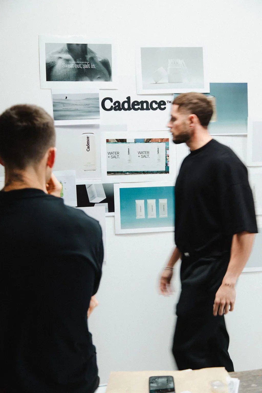 Two men are standing in front of a wall with multiple posters and branding materials for Cadence, including the words "WATER + SALT" and the Cadence logo.