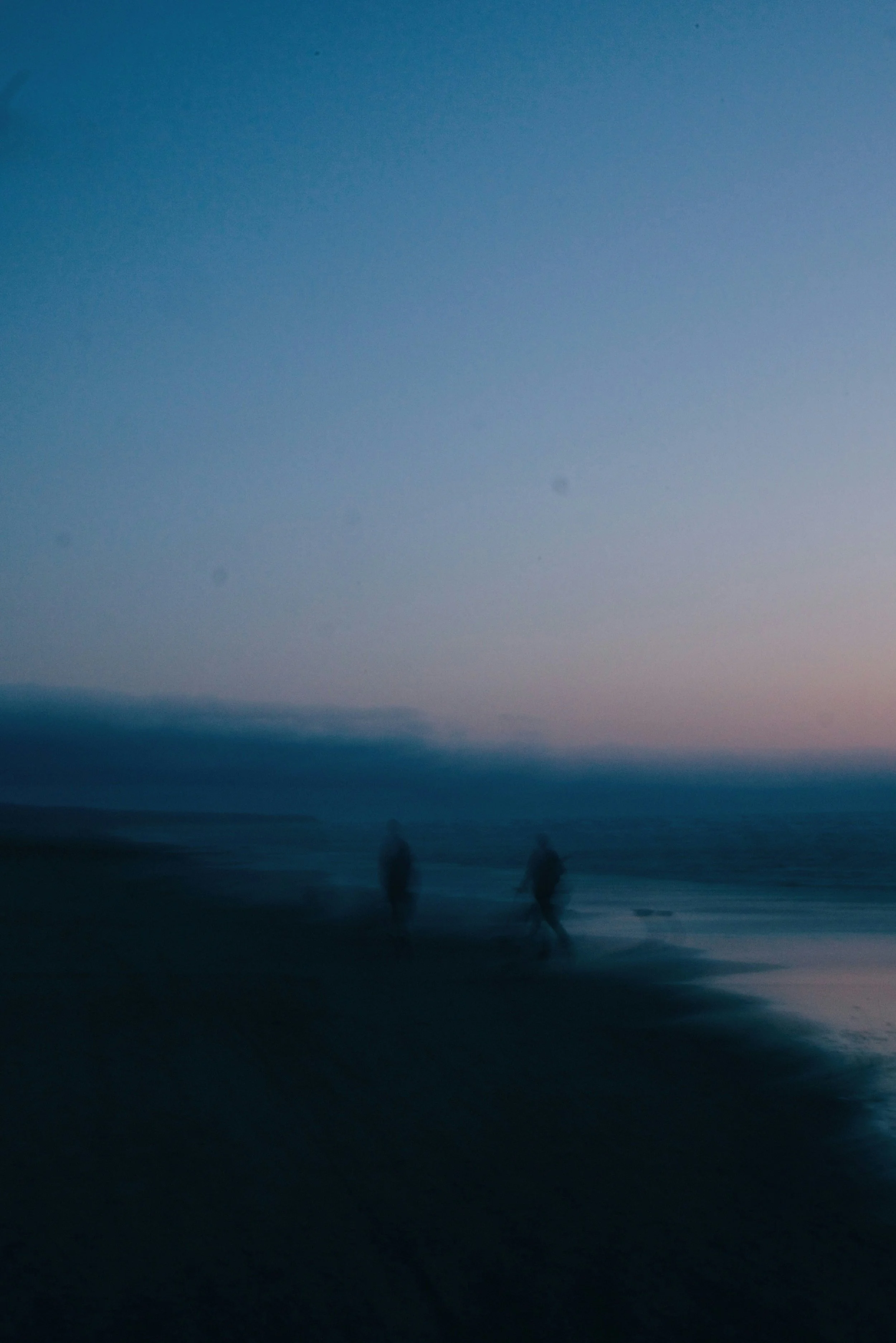Blurred image of two people walking along a beach at dusk with a cloudy sky and calm ocean waves.
