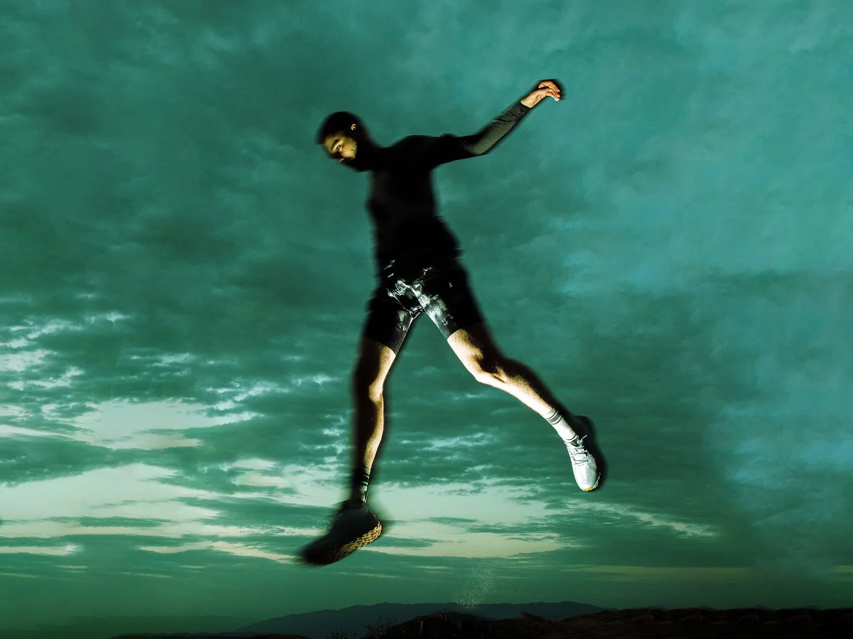 Person in athletic clothing jumping in the air outdoors during cloudy weather, with mountains visible in the background.